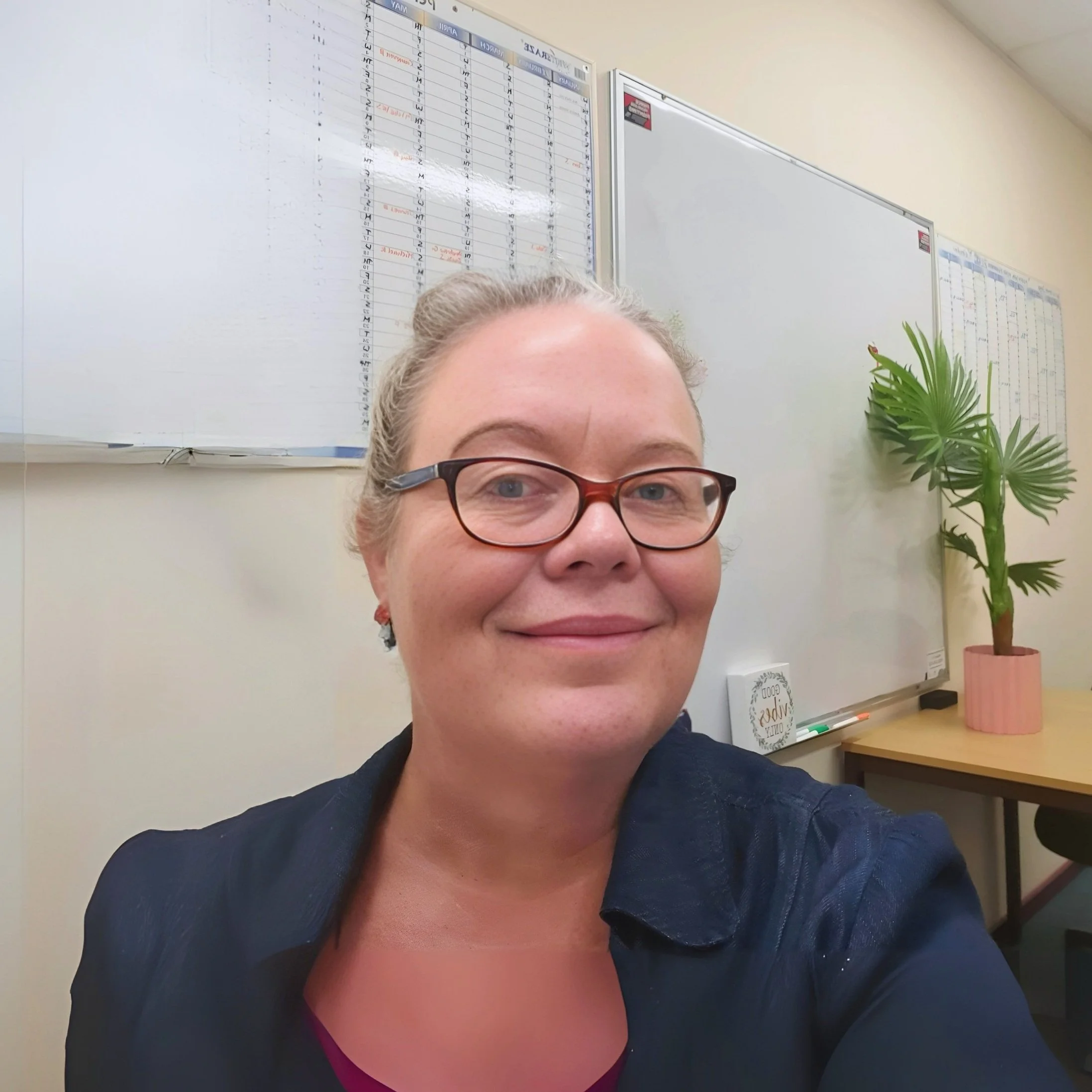 Image of Shannon, Support Coordinator A woman with glasses smiling in an office, with a whiteboard and a potted plant in the background.