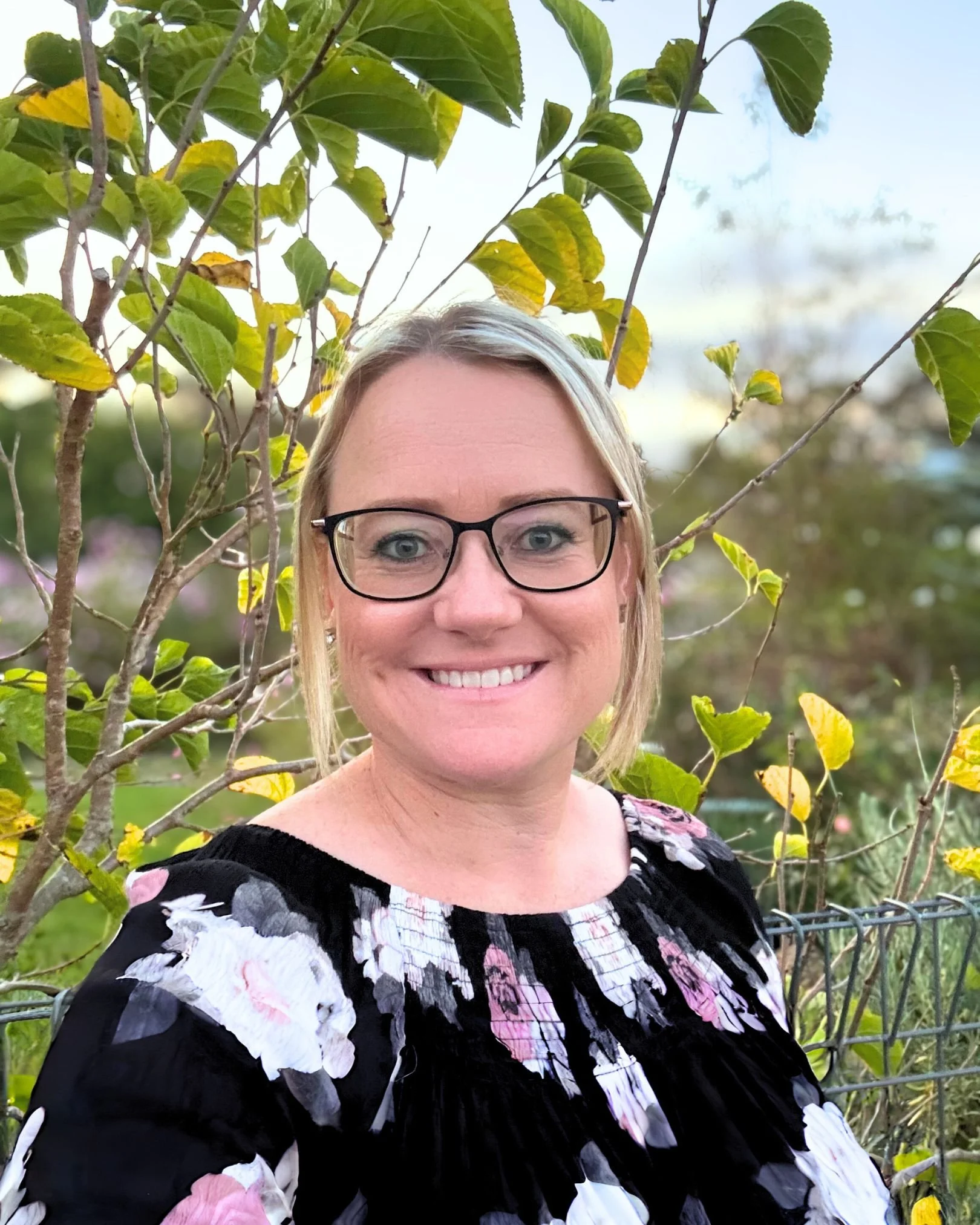 Image of Kristy, Senior Finance Officer - A smiling woman with blonde hair, glasses, and a red top standing outdoors with green trees in the background.