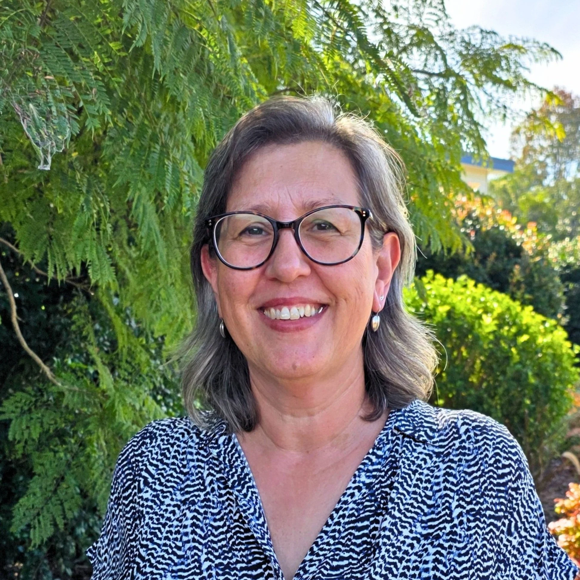Image of Alex, Finance Officer with glasses, shoulder-length gray hair, wearing earrings, smiling & standing outdoors in a lush green garden with trees.