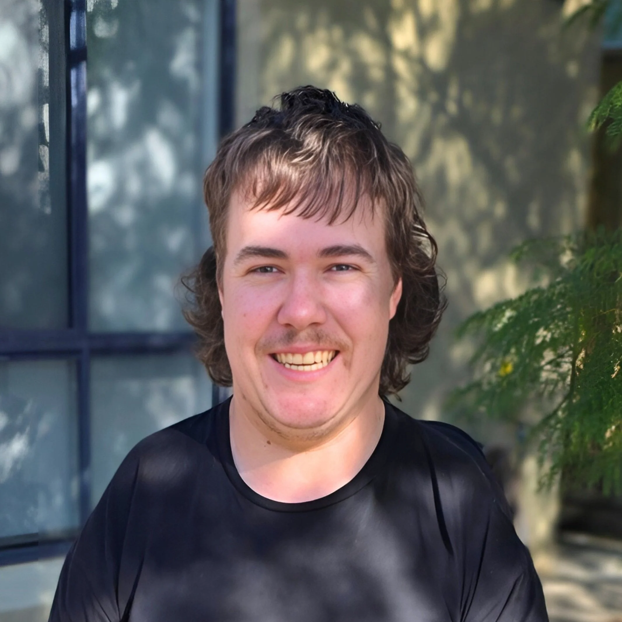 Image of Joel, Finance Officer - A young man with curly brown hair smiling while wearing a black shirt. Background includes a window and greenery.