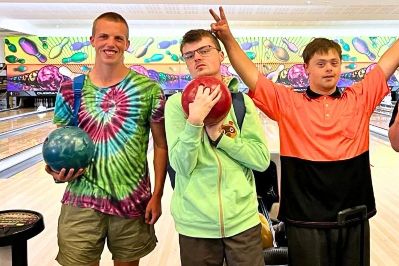 Four smiling members at a bowling alley, each holding a bowling ball, posing in front of the lanes