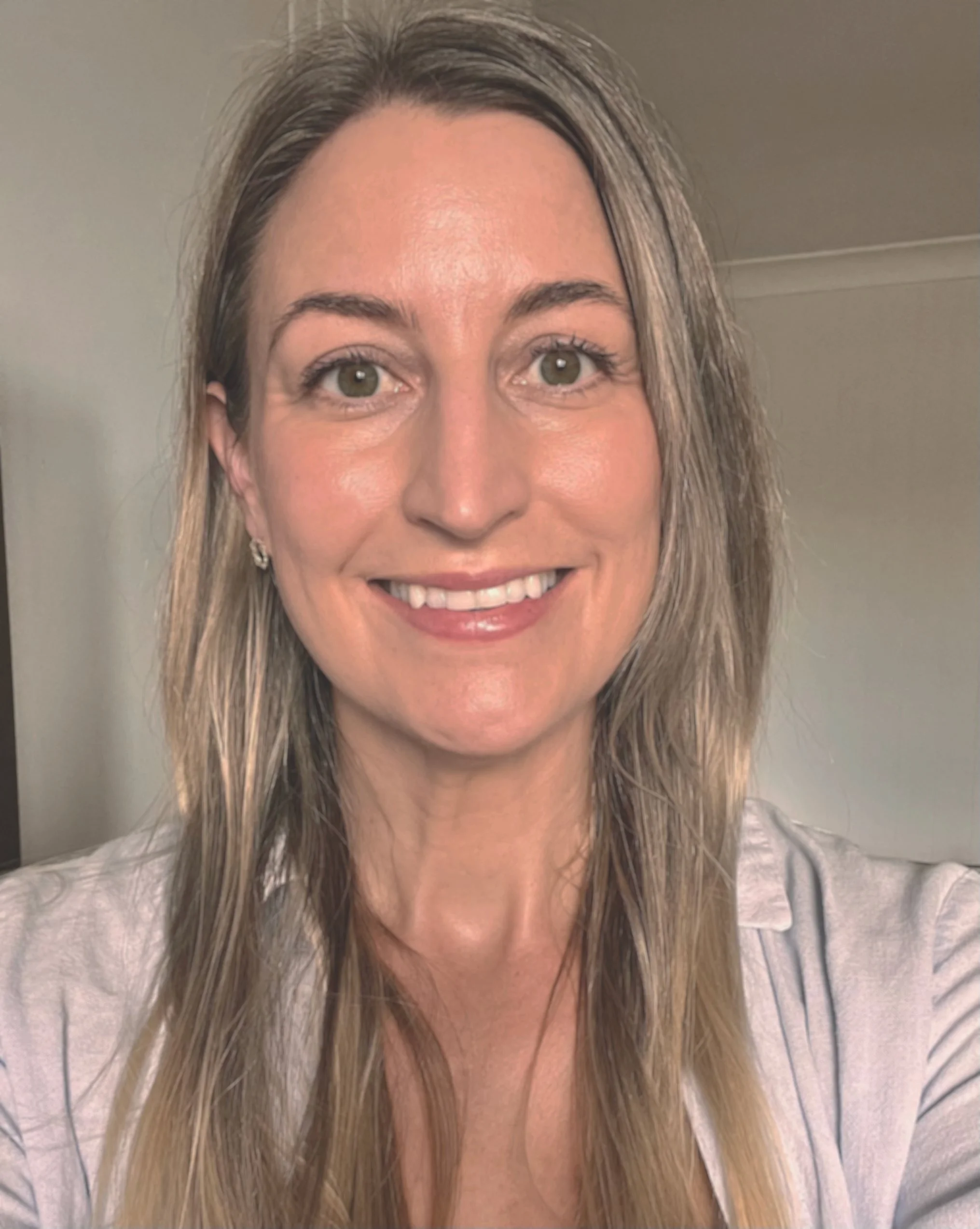 A woman smiling with shoulder-length blonde hair, wearing a light-colored top, indoors.