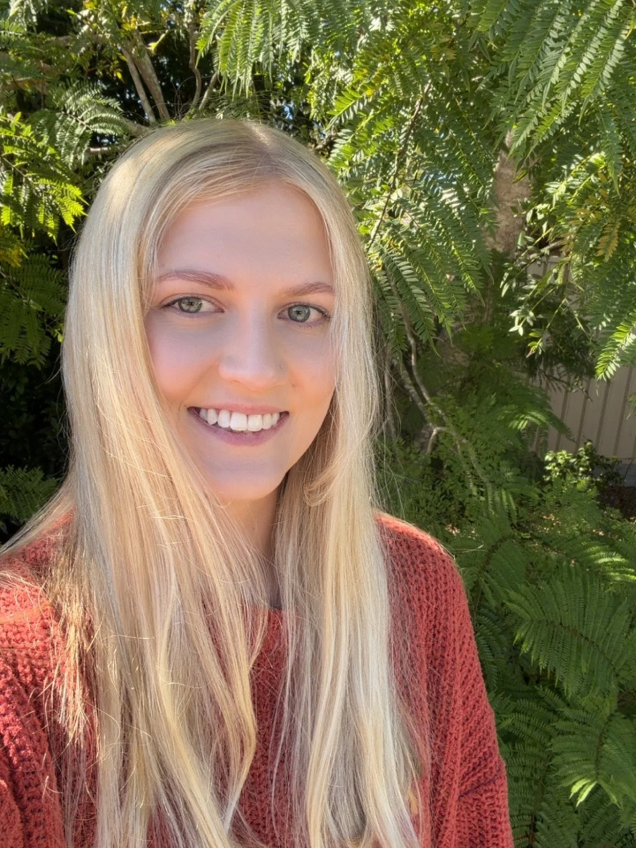 Image of Alana, Support Coordinator with shoulder-length blonde hair, blue eyes, and a subtle smile, wearing a white blouse with a red and black floral pattern, standing against a white paneled wall with decorative wooden branches.