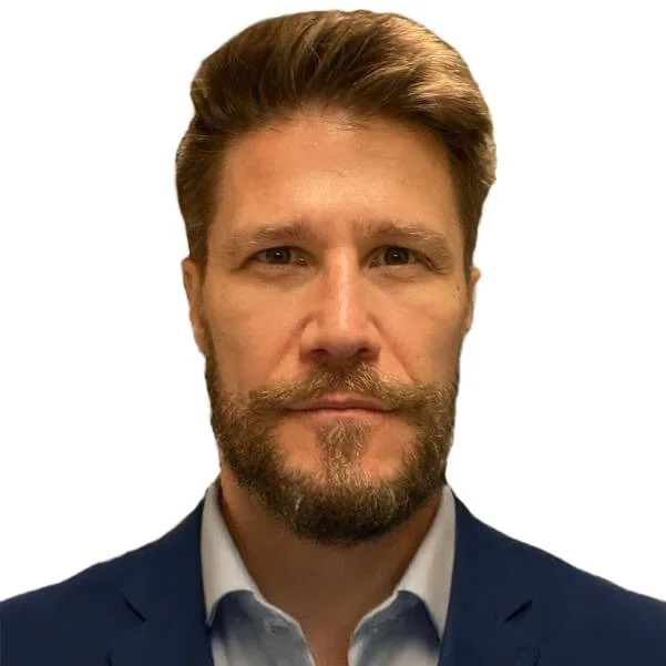 A professional headshot of a man with light skin, brown hair, a beard and mustache, wearing a blue suit and light blue shirt, against a white background.