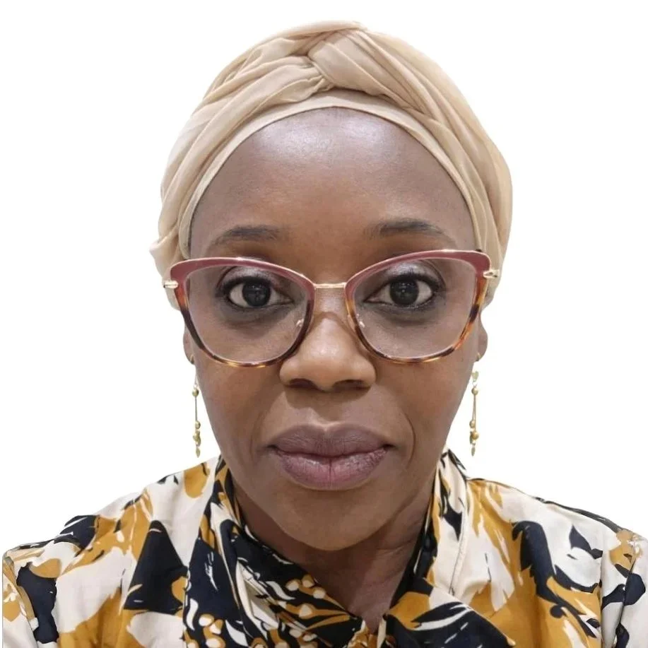A woman wearing a beige headwrap, pink eyeglasses, gold earrings, and a patterned shirt with black, white, and yellow colors.