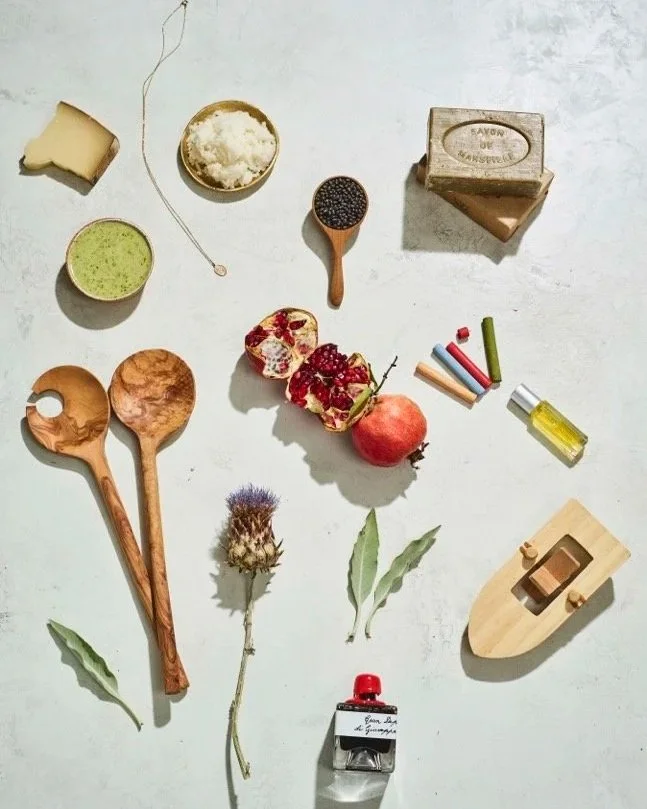 A flat lay of various objects including slices of bread, a bowl of green powder, a bowl of flour, a pomegranate, a pomegranate seed, a pomegranate flower, a bottle of cooking oil, a wooden spoon and spatula, a bar of soap, a necklace, chalks, and a small wooden toy boat, arranged on a white surface.