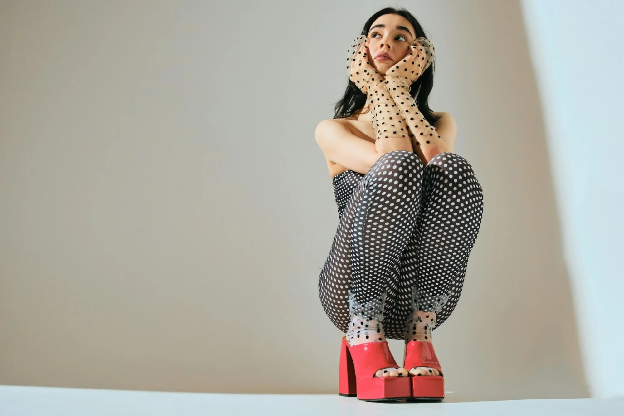 Nainen istuu polvillaan, jalat ristissä, punaiset platform-korkokengät, mustavalkoiset täplikkäät sukkahousut, pitsiset käsineet, jotka peittävät kasvot.
