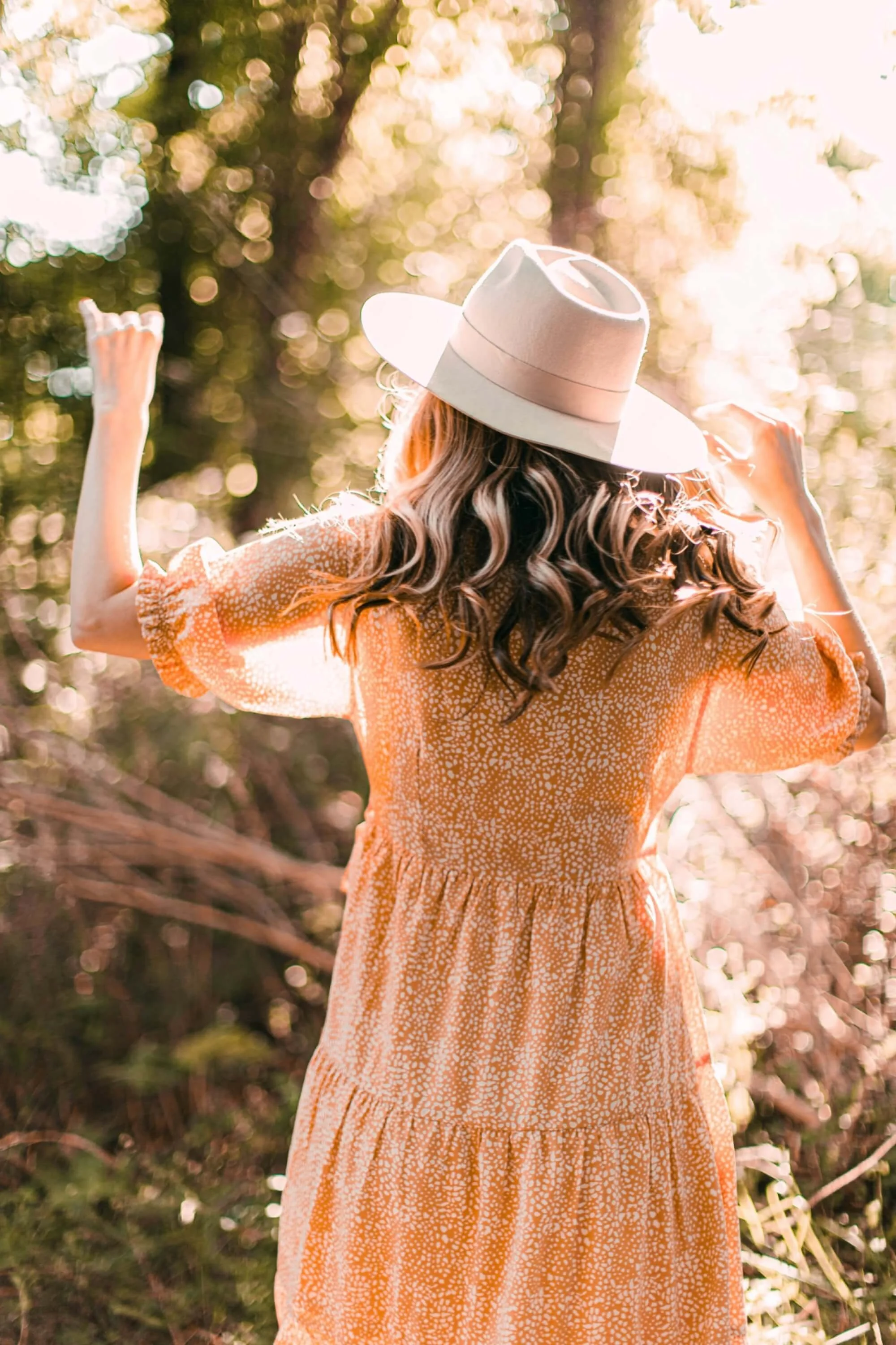 Nainen, jolla on pitkä, kihara tukka, katsoo kauas ja pitää käsiään ylös metsän keskellä, auringonvalossa.