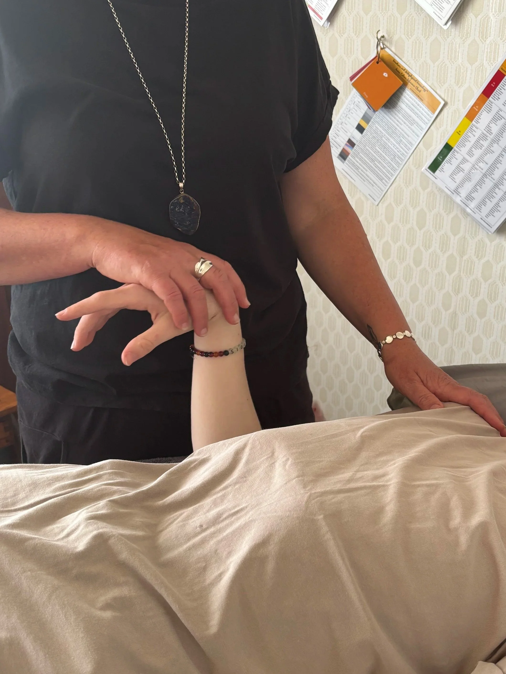 A person is holding a child's hand in a hospital room. The person wears jewelry including a long necklace, a ring, and bracelets. The child's hand has a bracelet. The background shows informational posters on a wall.