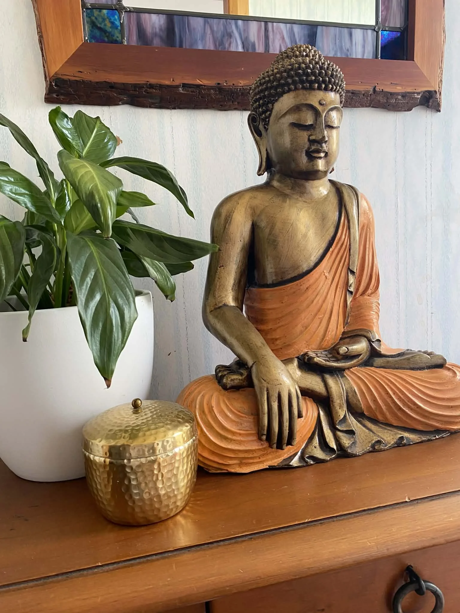 Decorative wooden Buddha statue with closed eyes, orange robe, seated on a wooden surface next to a green potted plant and a gold textured container.