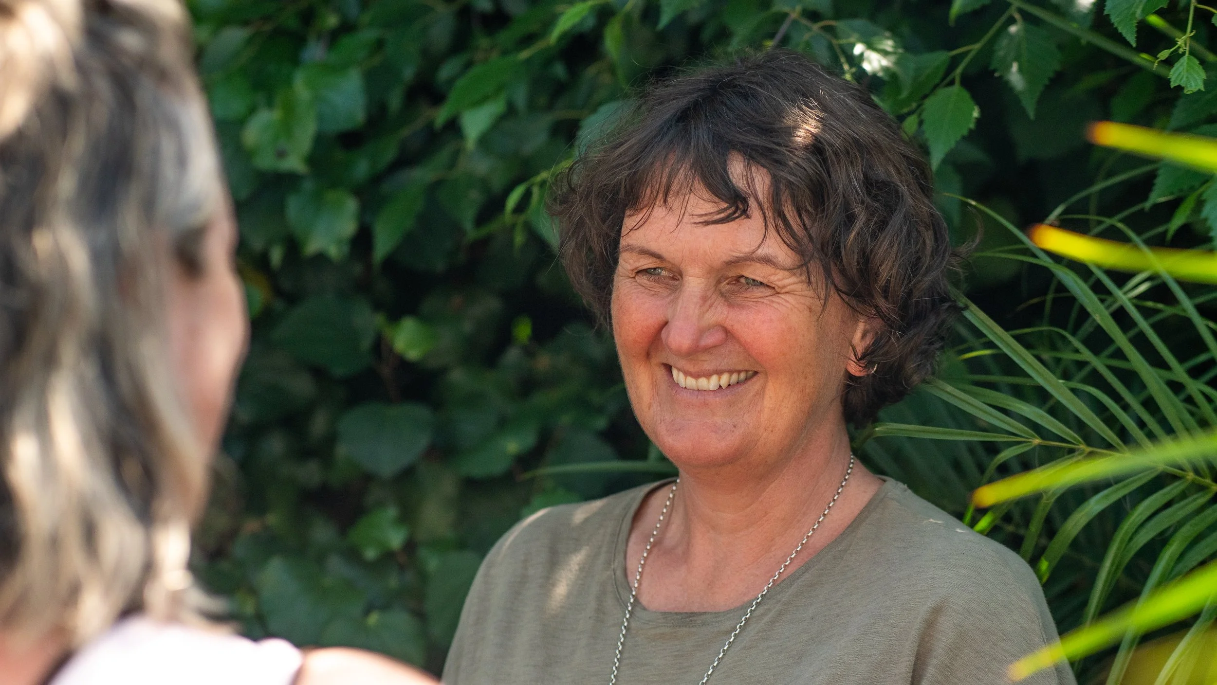 A woman smiling outdoors, engaging in conversation with another person whose face is partially visible, surrounded by green foliage.