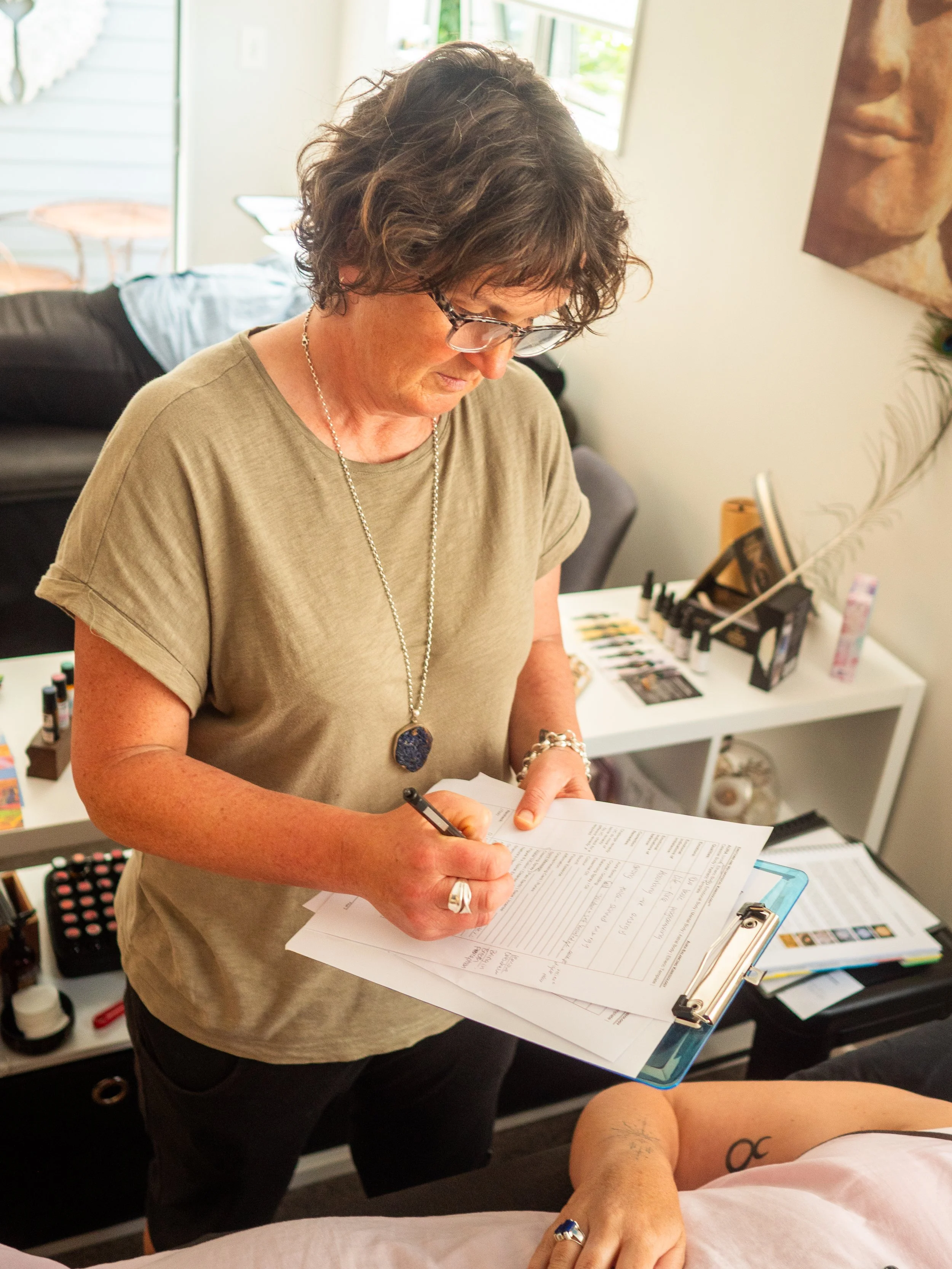 A woman with curly hair and glasses writing on papers at a desk during a tattoo appointment, with a tattooed person's arm in the foreground.
