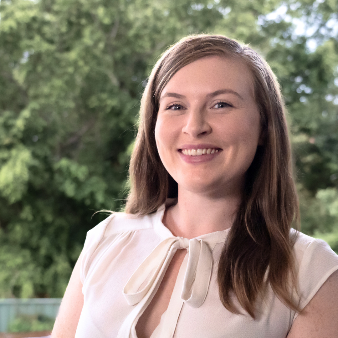 A young woman with light skin and long brown hair smiling outdoors with trees in the background.