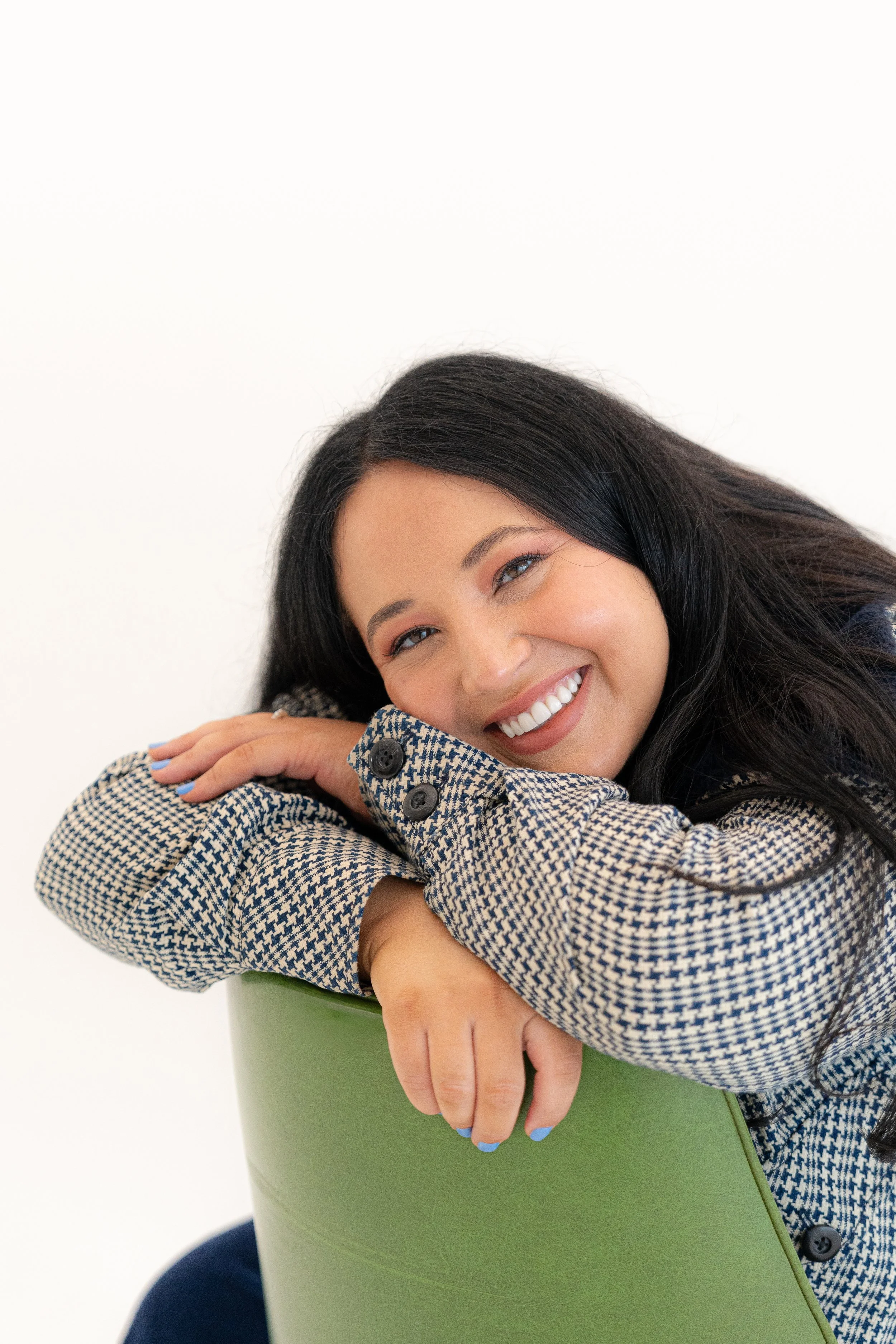 Close-up of a woman with long black hair smiling, resting her head on her crossed arms on the back of a green chair, wearing a houndstooth patterned jacket