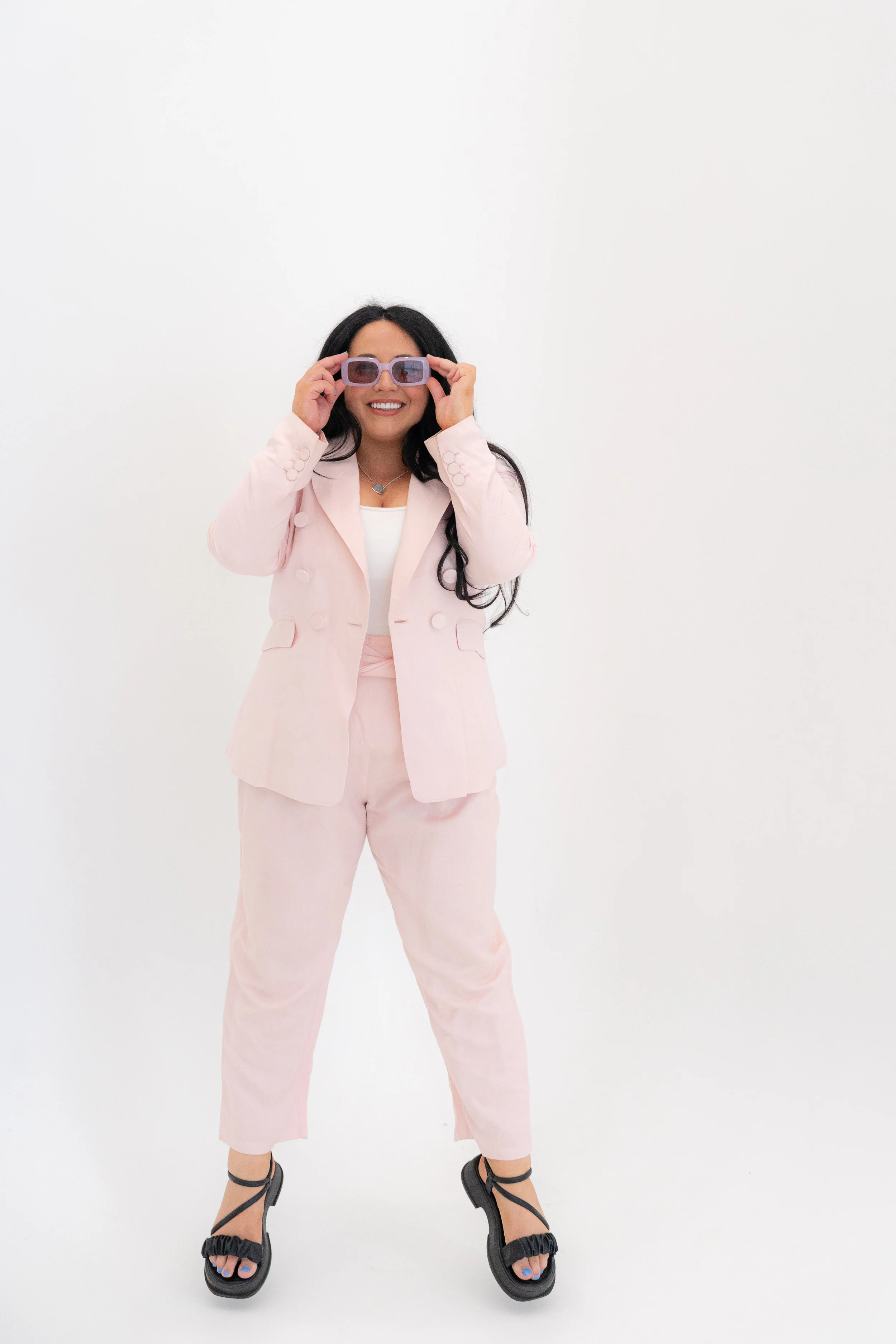 A woman in a pink suit holding purple sunglasses, smiling against a white background.