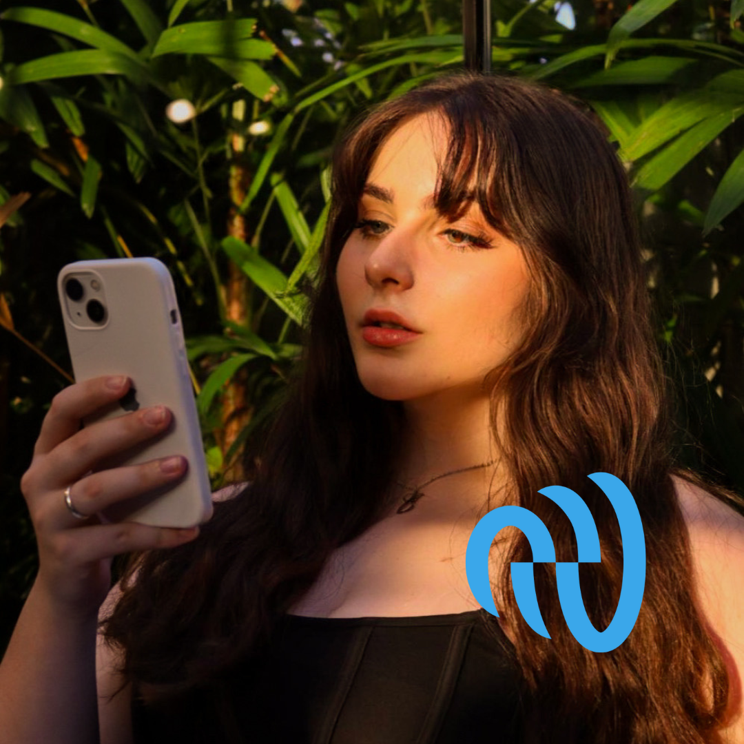 Young woman with long brown hair and fair skin looking at a smartphone in a lush green outdoor setting.