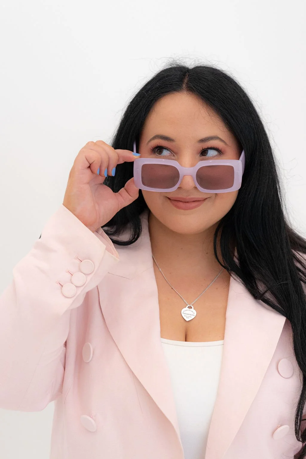 A woman with long black hair and a light pink blazer is holding sunglasses, revealing her eyes. She is smiling slightly and appears to be looking to her left.