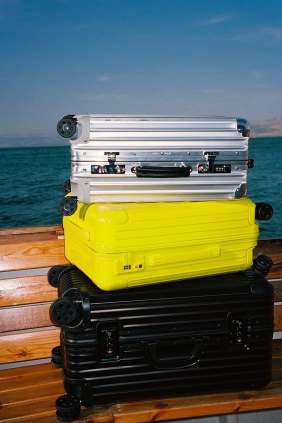 Four hard-shell suitcases stacked on a wooden surface outdoors near water, with a clear sky in the background.