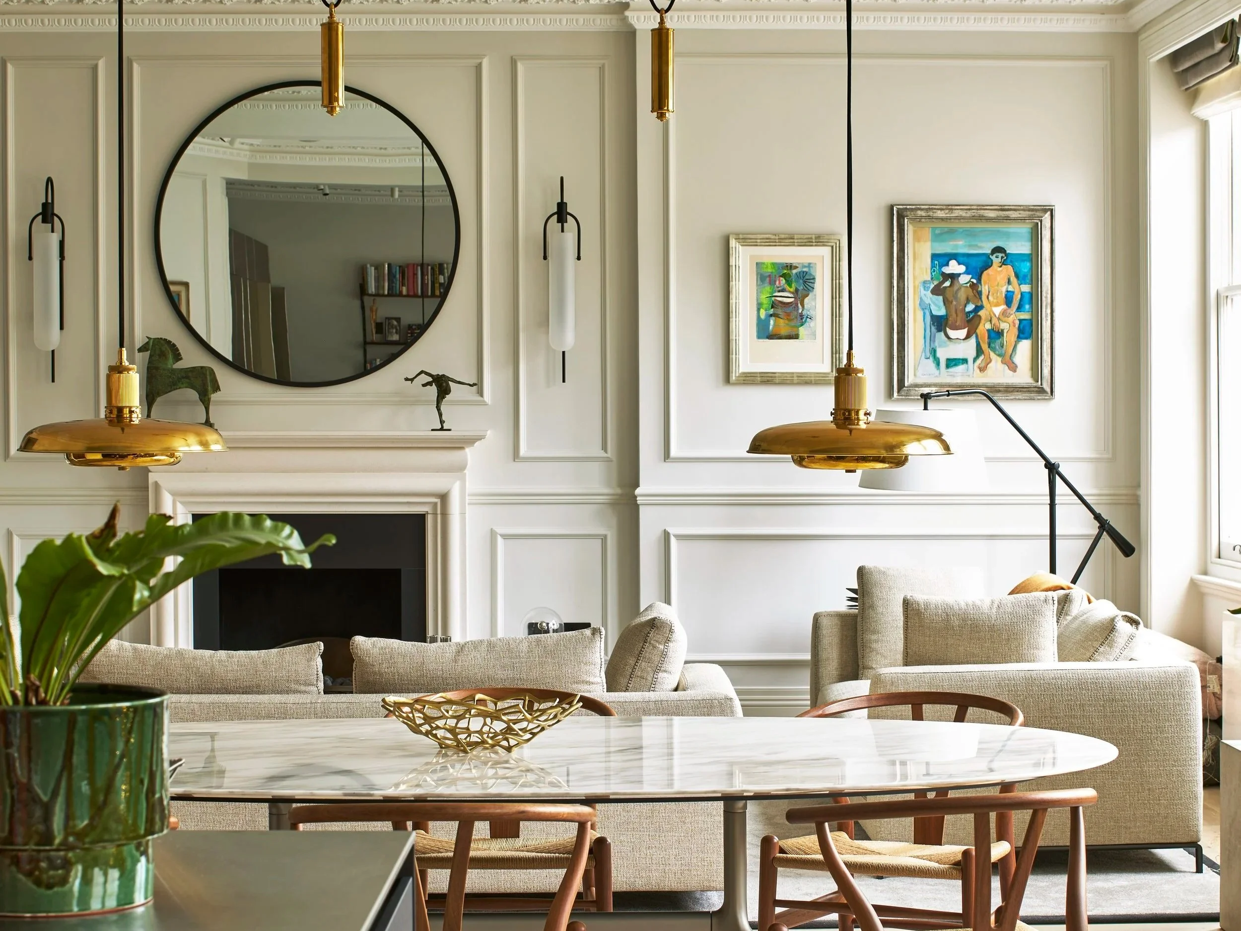Living room with white wall paneling, three framed paintings, large window, beige sofas, wooden chairs, marble dining table with a green potted plant, gold pendant lights, mirror above fireplace, sculptures, and a black floor lamp.