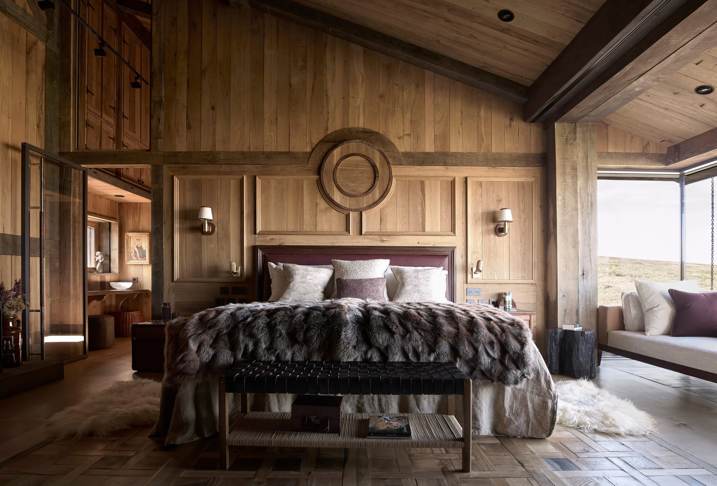 A bedroom with wooden walls, ceiling, and floors, featuring a large bed with multiple pillows and a fur throw, a bench at the foot of the bed, and a window on the right showing an outdoor landscape.