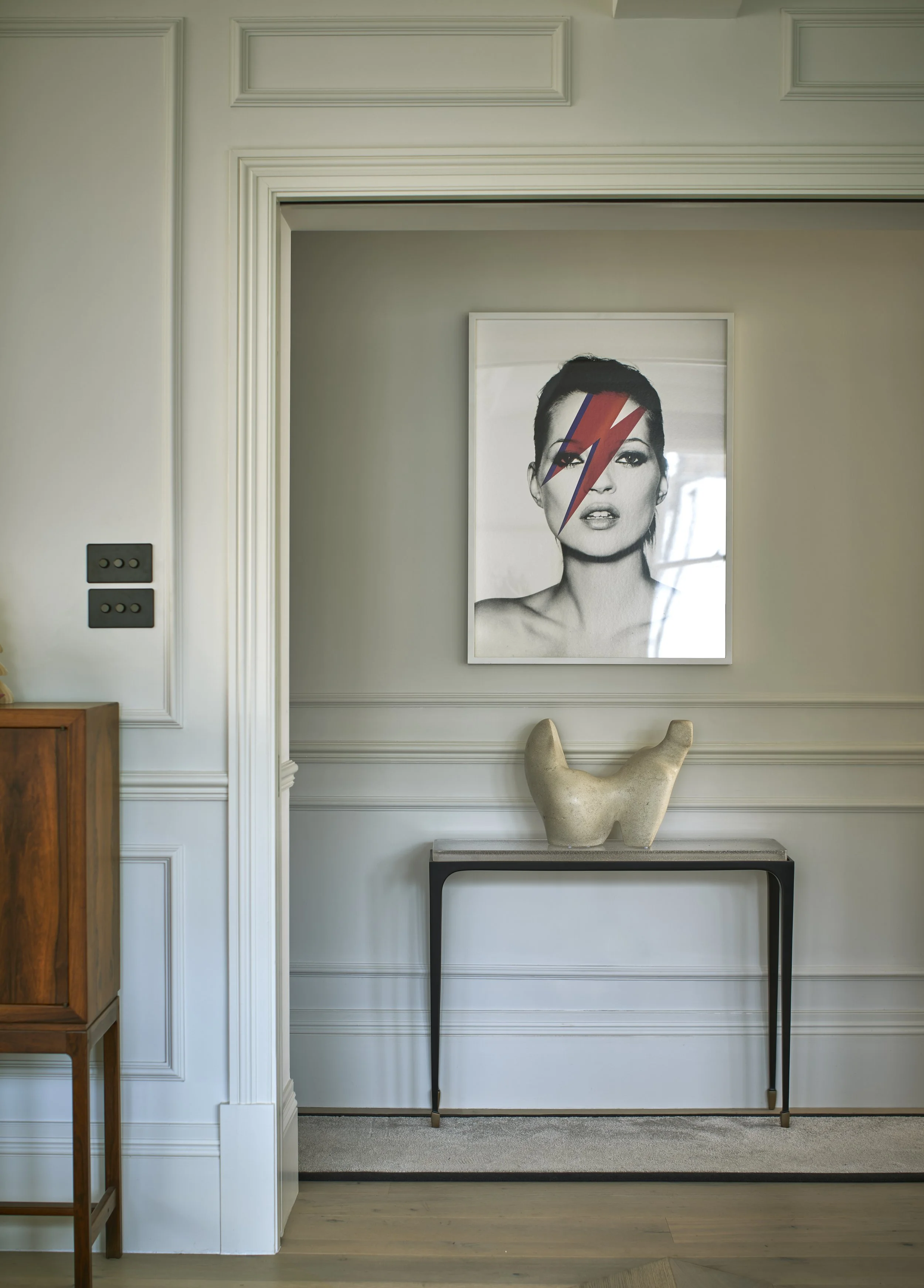 A black and white photograph of a woman with red lipstick, featuring a colorful lightning bolt face graphic, hanging on a white wall above a black console table with a decorative sculpture. The room has white paneled walls and light hardwood flooring.