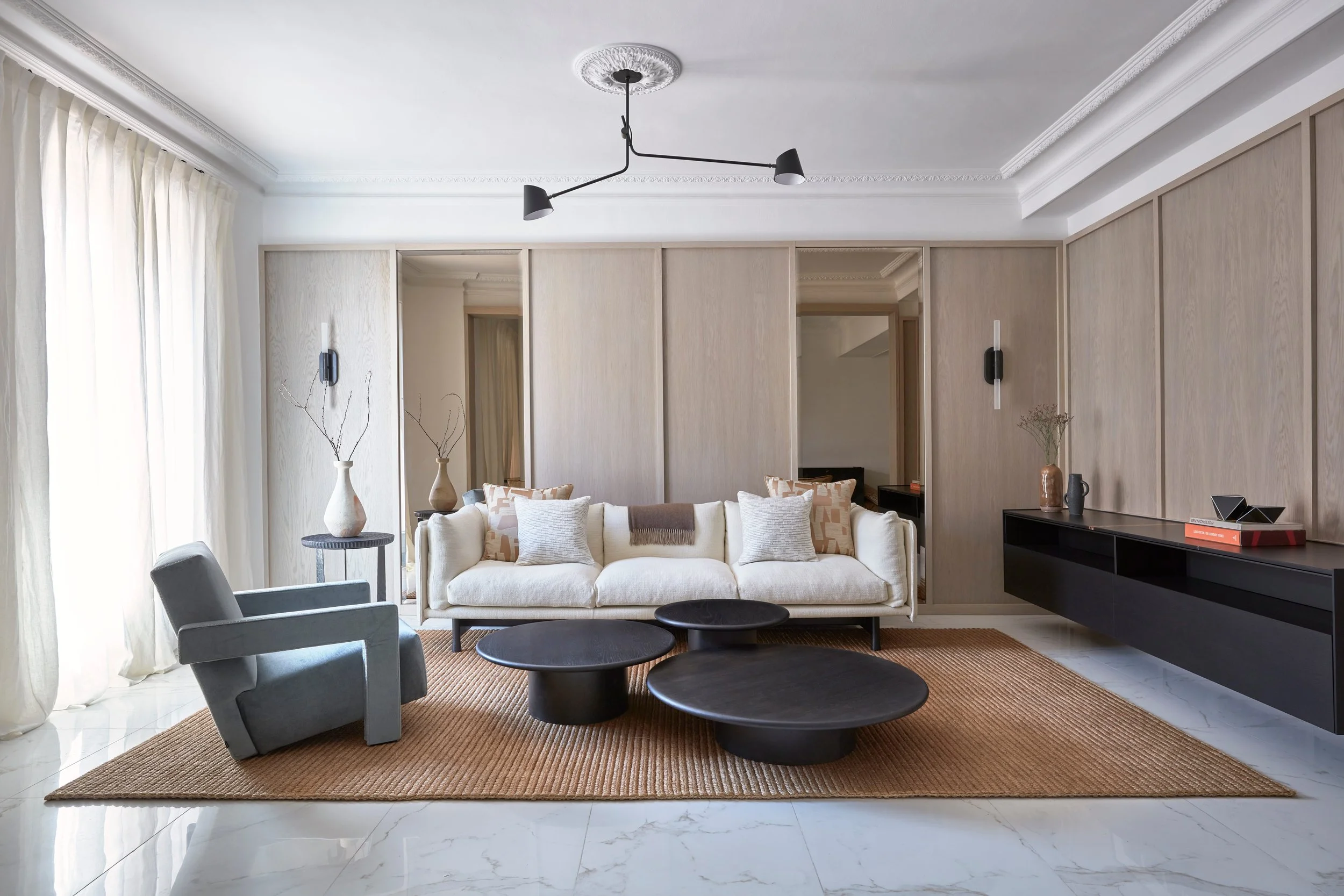 Modern living room with white sofa, gray armchair, black coffee tables, beige rug, black wall-mounted console, and decorative vases with branches. Large window with white curtains, light wood-paneled wall, and ceiling light fixture.