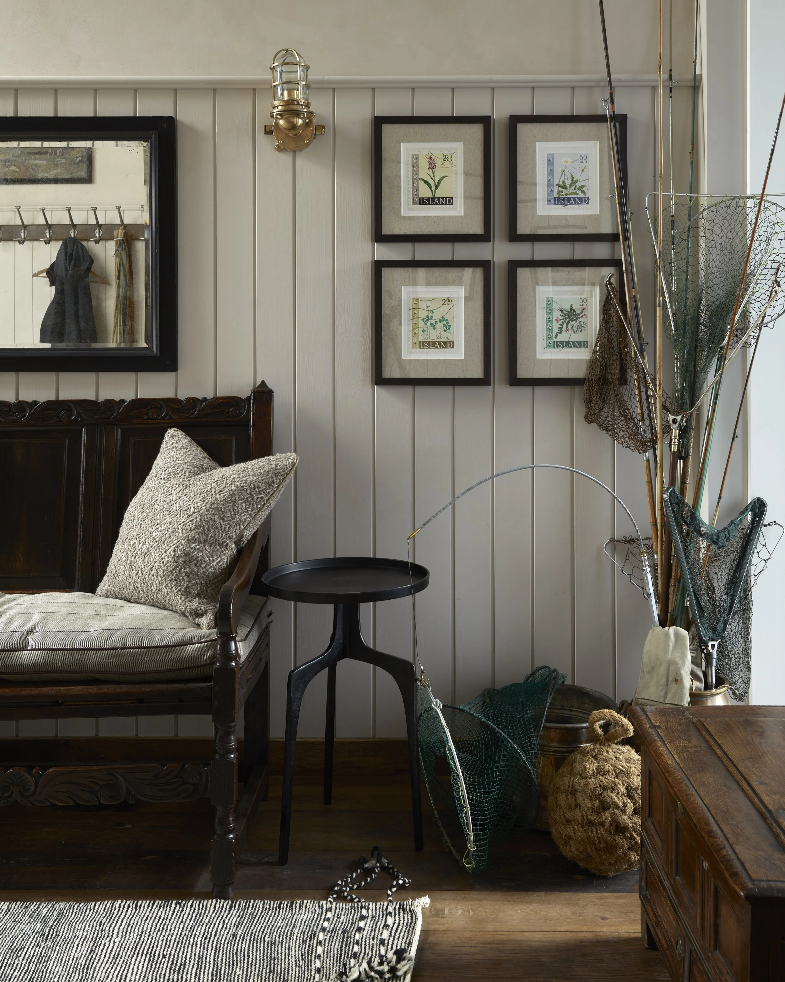 Cozy interior with wooden bed and armchair, framed botanical prints on wall, fishing gear and netting stored in corner, and mirror reflection of hanging clothes.