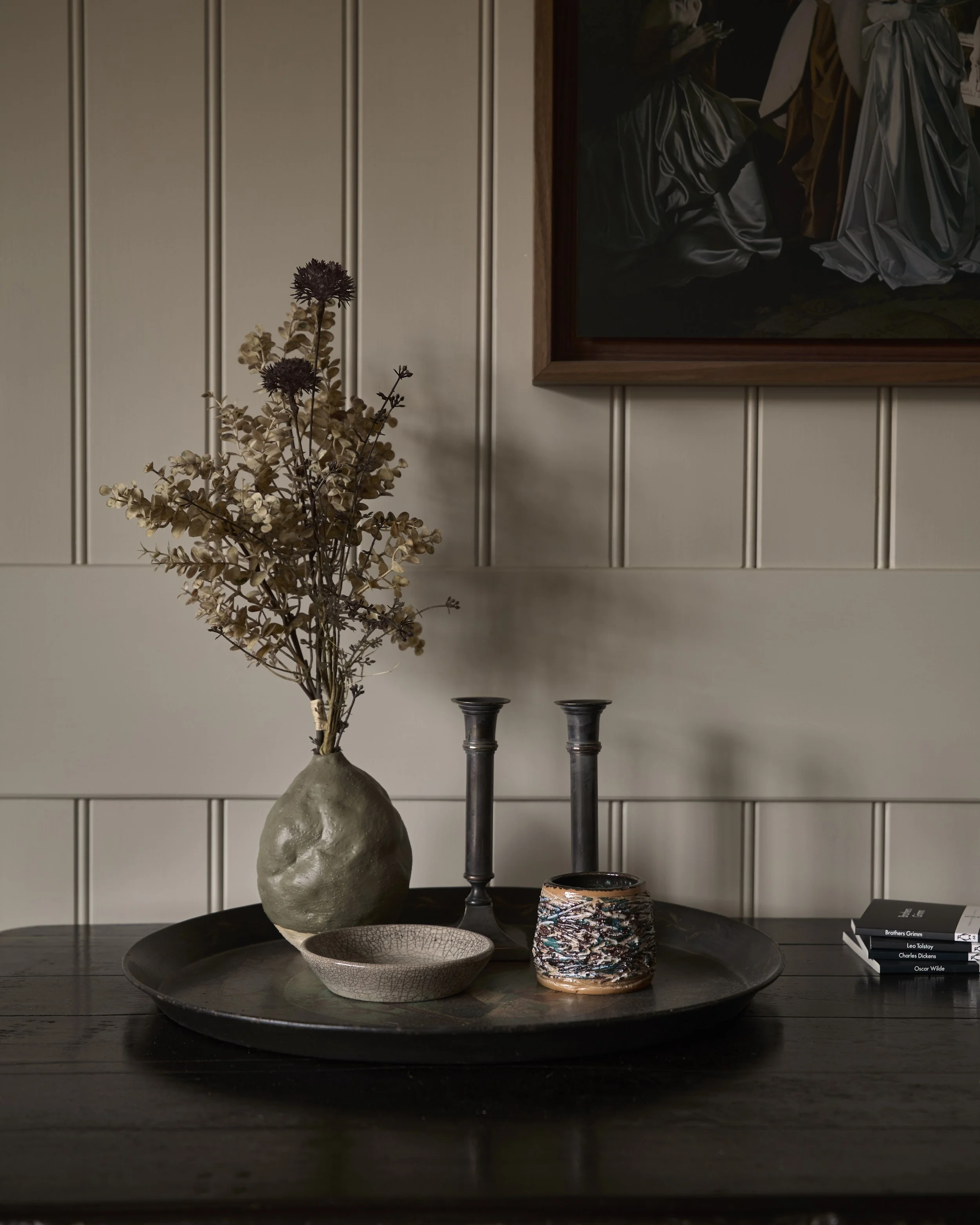 Decorative table with a large vase holding dried flowers, two black candlesticks, a textured bowl, a painted ceramic pot, and a small stack of books, against a paneled wall with a framed painting.