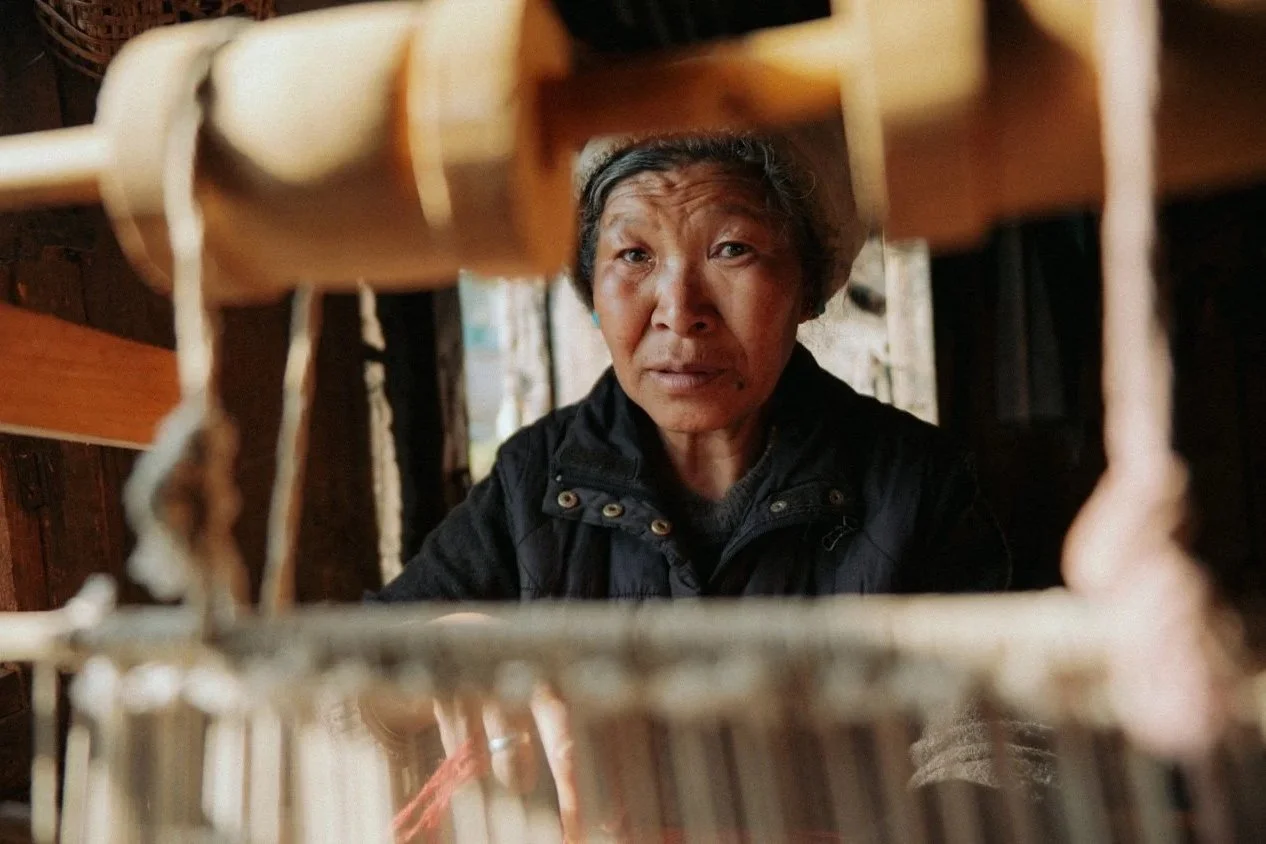 Eine ältere asiatische Frau schaut durch eine kleine offene Tür oder ein Fenster in eine Kamera, mit einem ernsten Ausdruck auf dem Gesicht.