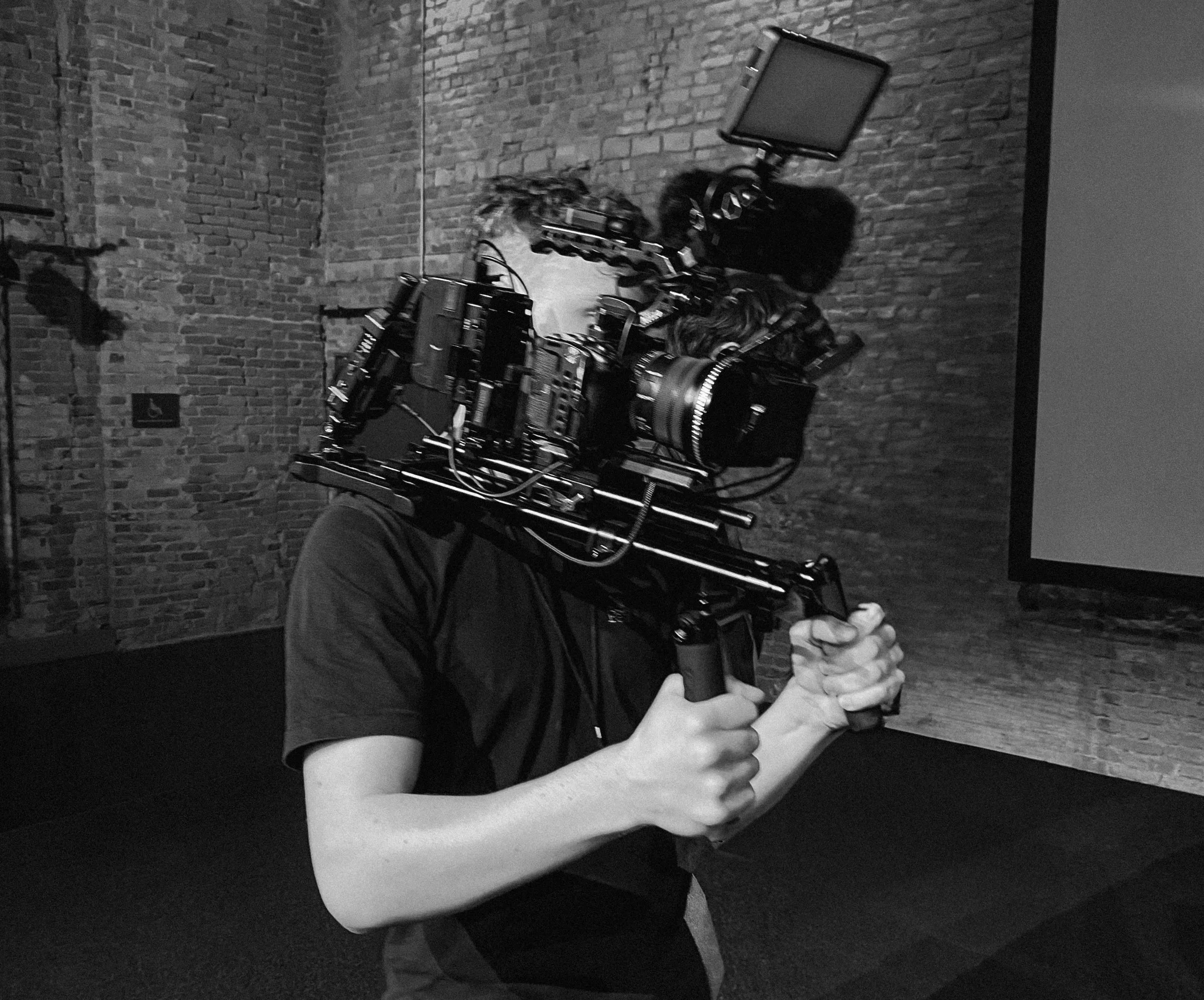 Ein Mensch hält eine große Filmkamera vor seinem Gesicht, was die Sicht auf sein Gesicht verdeckt. Der Hintergrund besteht aus einer Ziegelmauer.