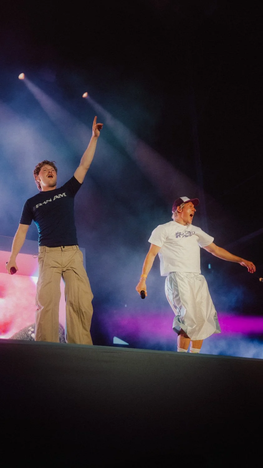 Zwei Männer auf einer Bühne, einer mit erhobener Hand und das andere mit Mikrofon, bei einer Studio- oder Bühnenveranstaltung mit buntem Licht.