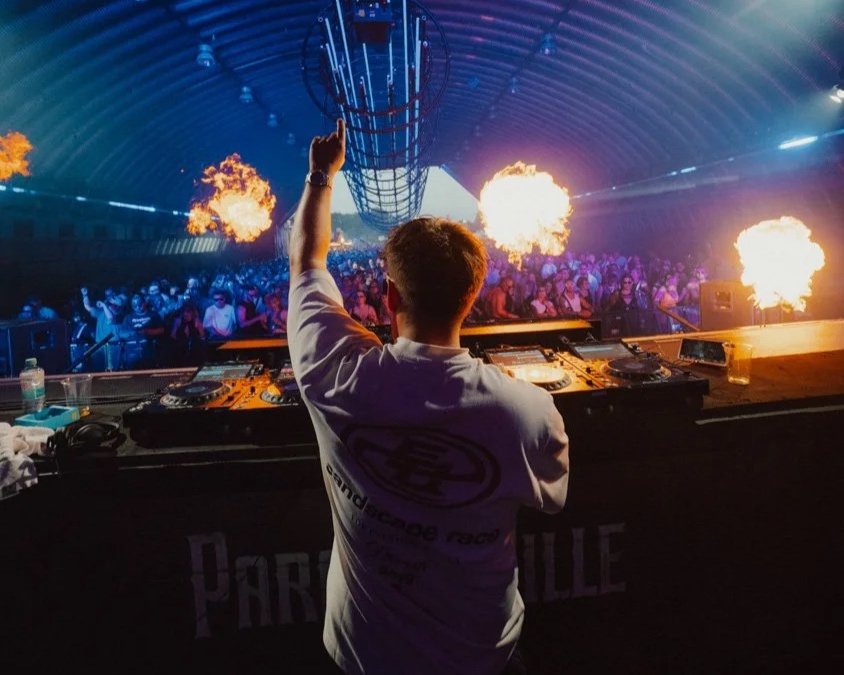 DJ auf der Bühne mit brennenden Flammen, Blick auf die tanzende Menge in einem Club mit farbigem Licht.