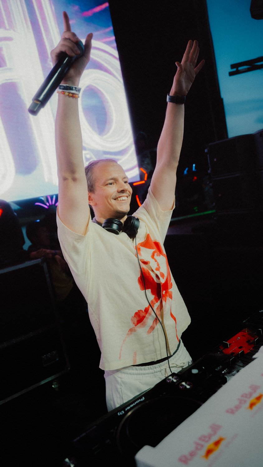 Ein DJ bei einer Party, der begeistert in die Luft streckt, mit Kopfhörern um den Hals und einem Mikrofon in der Hand, im Hintergrund bunte Lichter und eine große Leinwand mit Neon-Design.