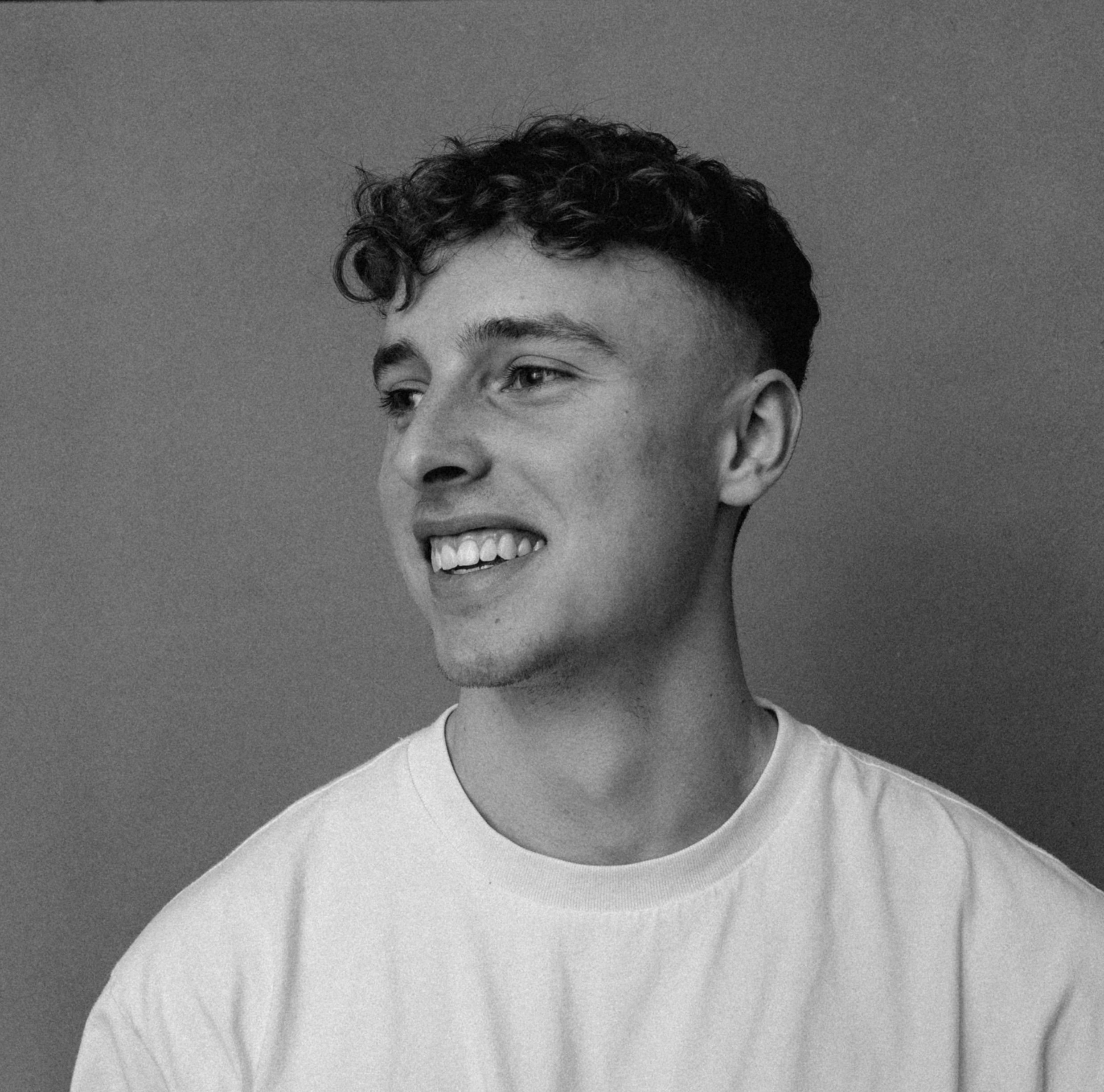 Schwarz-Weiß-Foto eines jungen Mannes, der nach rechts lächelt, mit kurzen, lockigen Haaren, weißem T-Shirt und hellem Hintergrund.