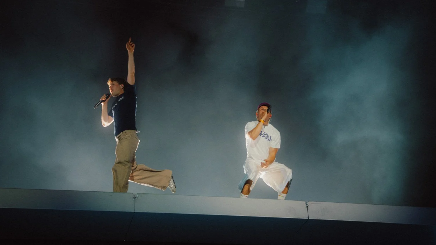 Zwei junge Männer singen auf einer Bühne, beide tragen Mikrofone und befinden sich in dynamischer Pose, mit dunklem, rauchigem Hintergrund.