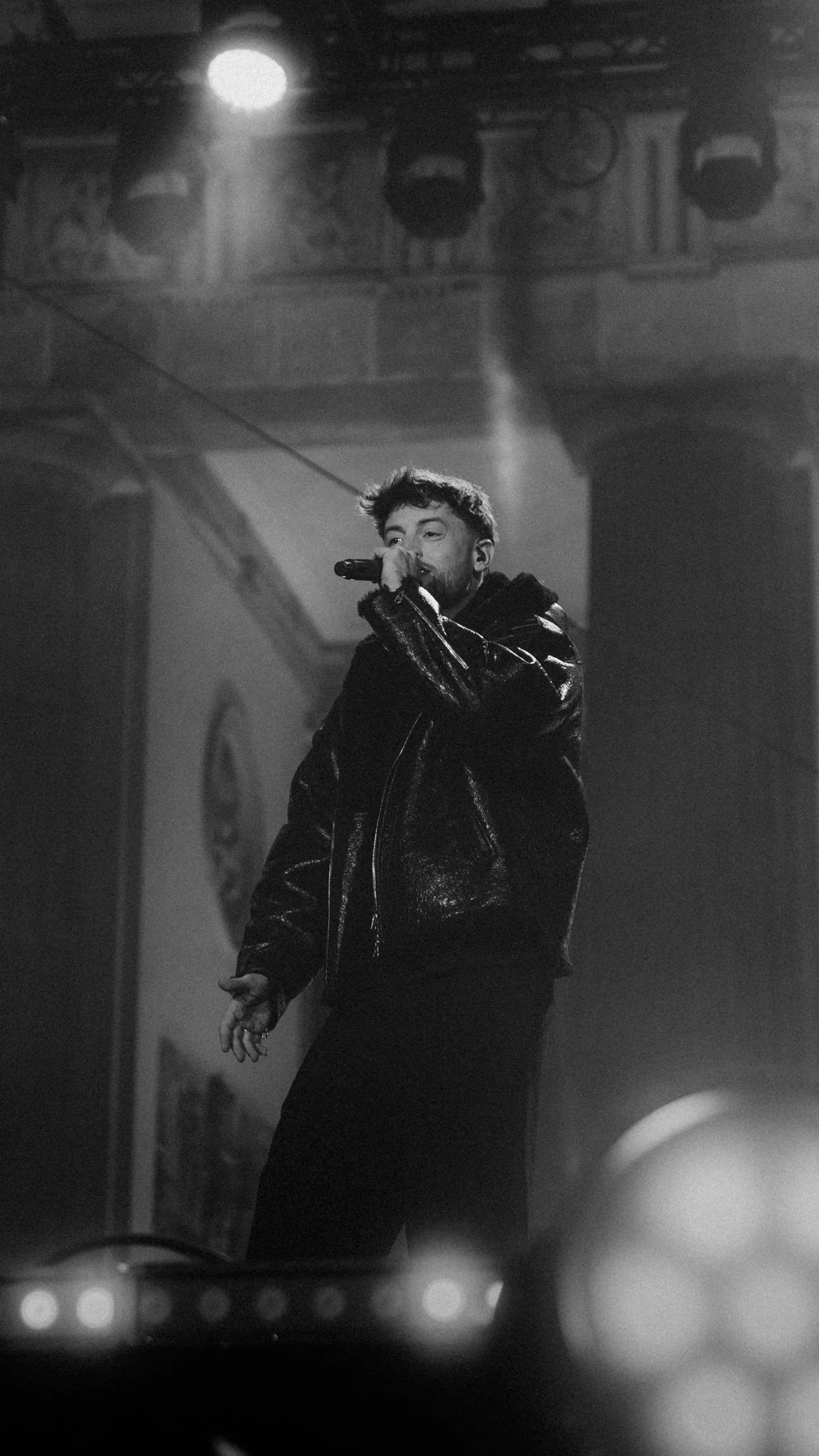 Mann singt auf einer Bühne mit einem Mikrofon, schwarze Lederjacke, im Schwarzweißfoto, mit Scheinwerferlicht im Hintergrund.