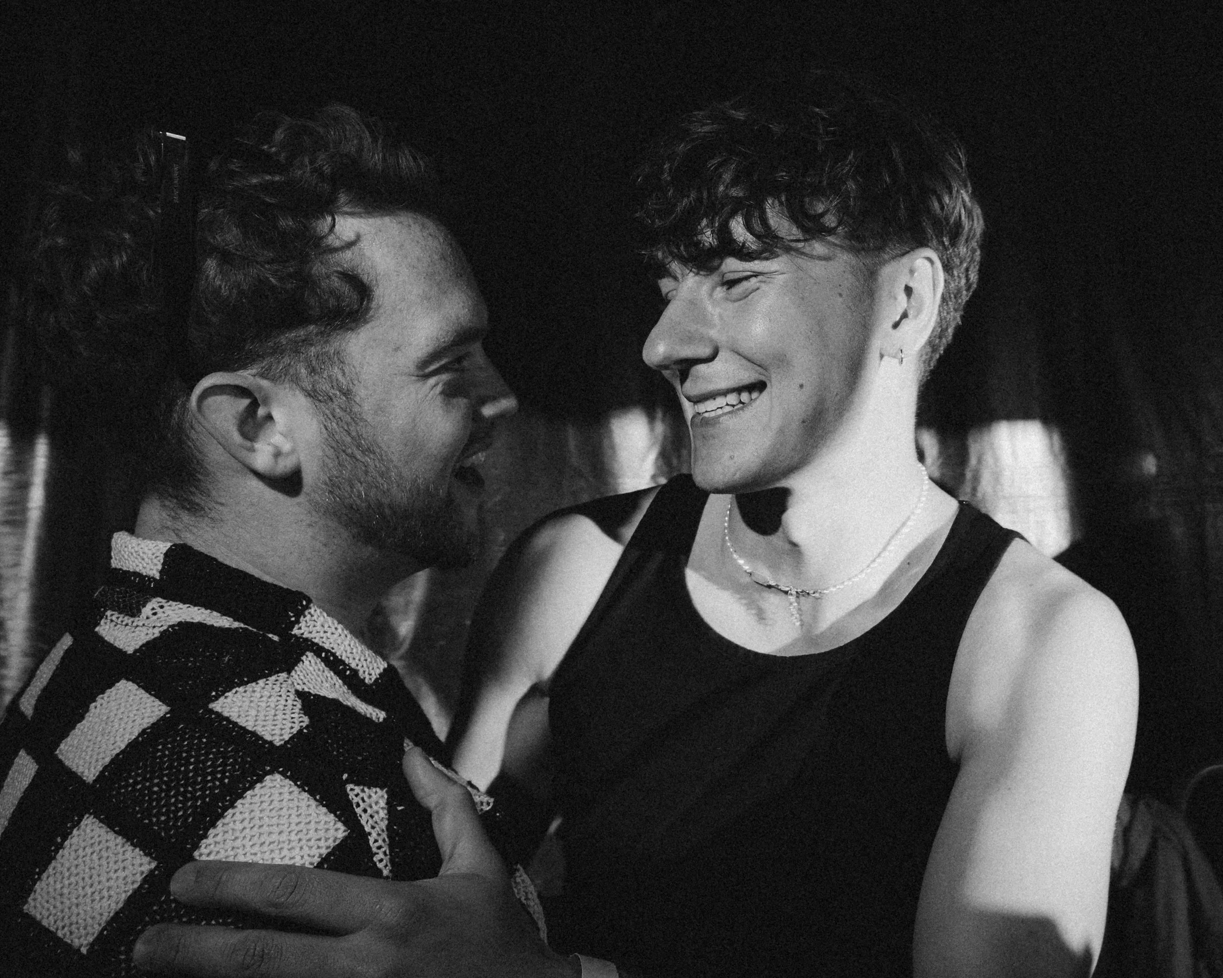 Zwei lachende Menschen, ein Mann mit Bart und lockigen Haaren, eine Frau mit kurzen, lockigen Haaren, die sich in einem dunklen Raum umarmen und lachen.