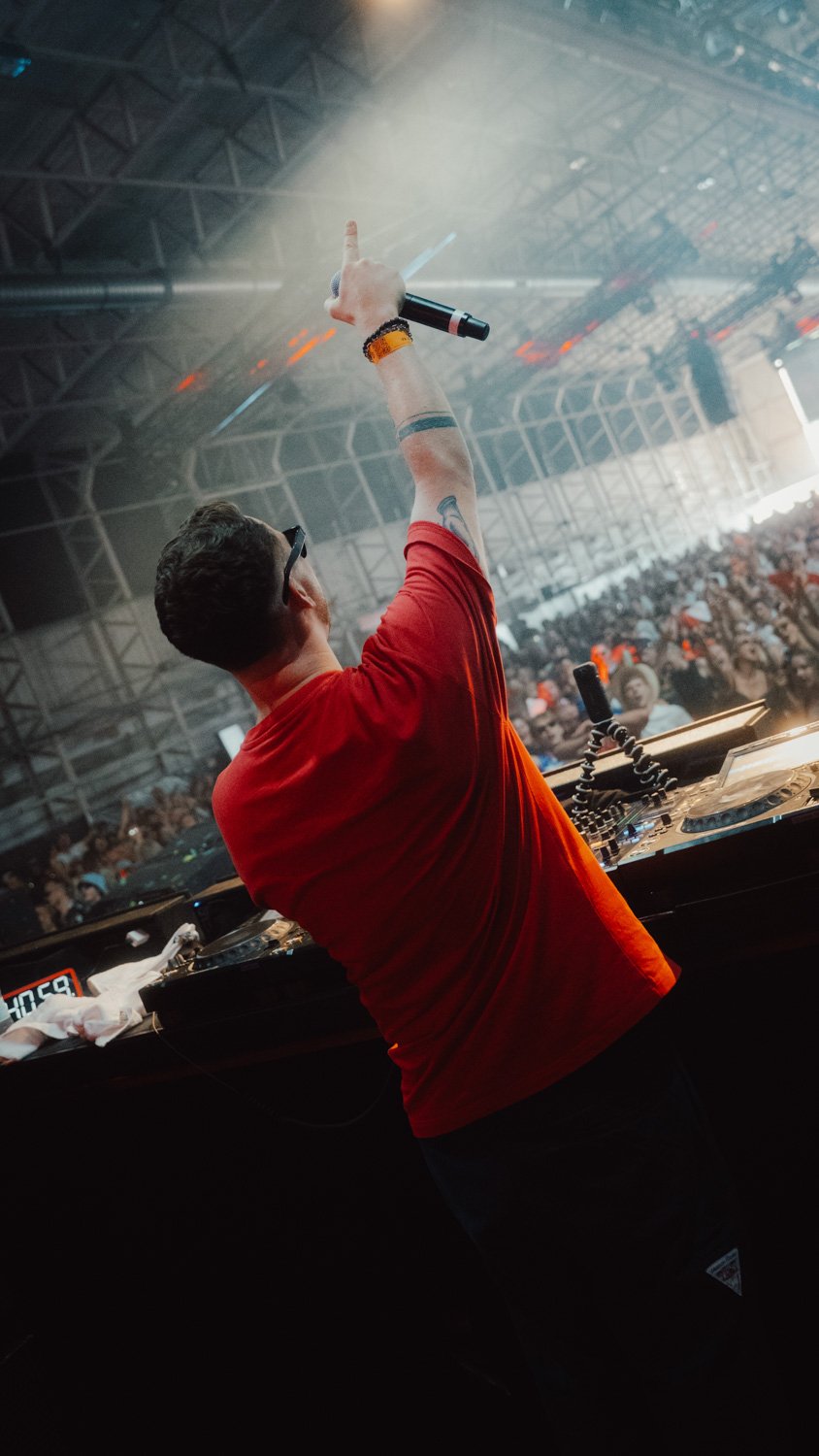 DJ auf der Bühne in einem großen Indoor-Konzertsaal, schaut auf die Crowd und hebt die Hand mit einem Mikrofon empor, um die Menge zu animieren.