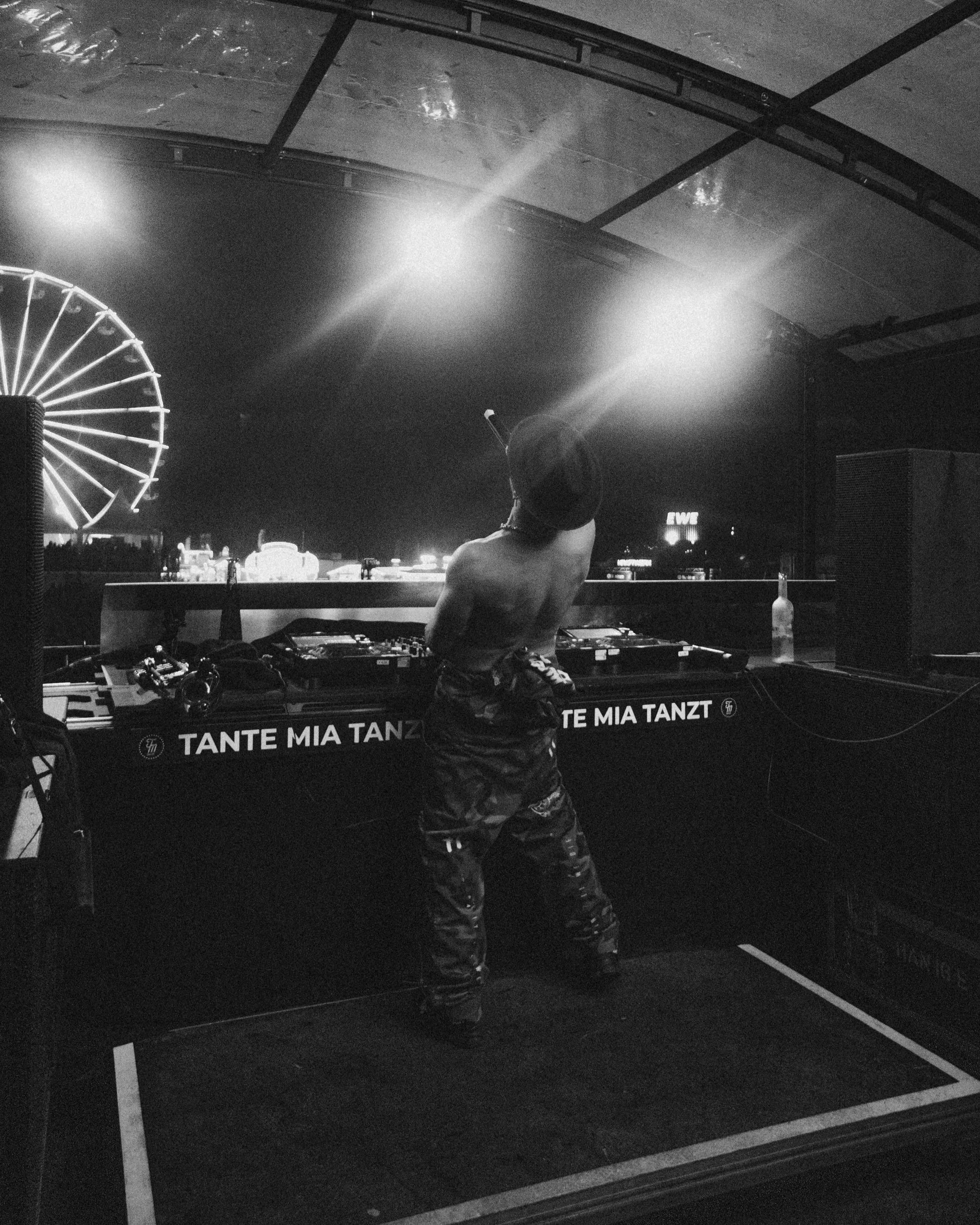 Eine Person steht hinter einem DJ-Pult im Zelt, bei Nacht. Es gibt Lichter und ein Riesenrad im Hintergrund, es scheint eine Feier oder ein Festival zu sein.