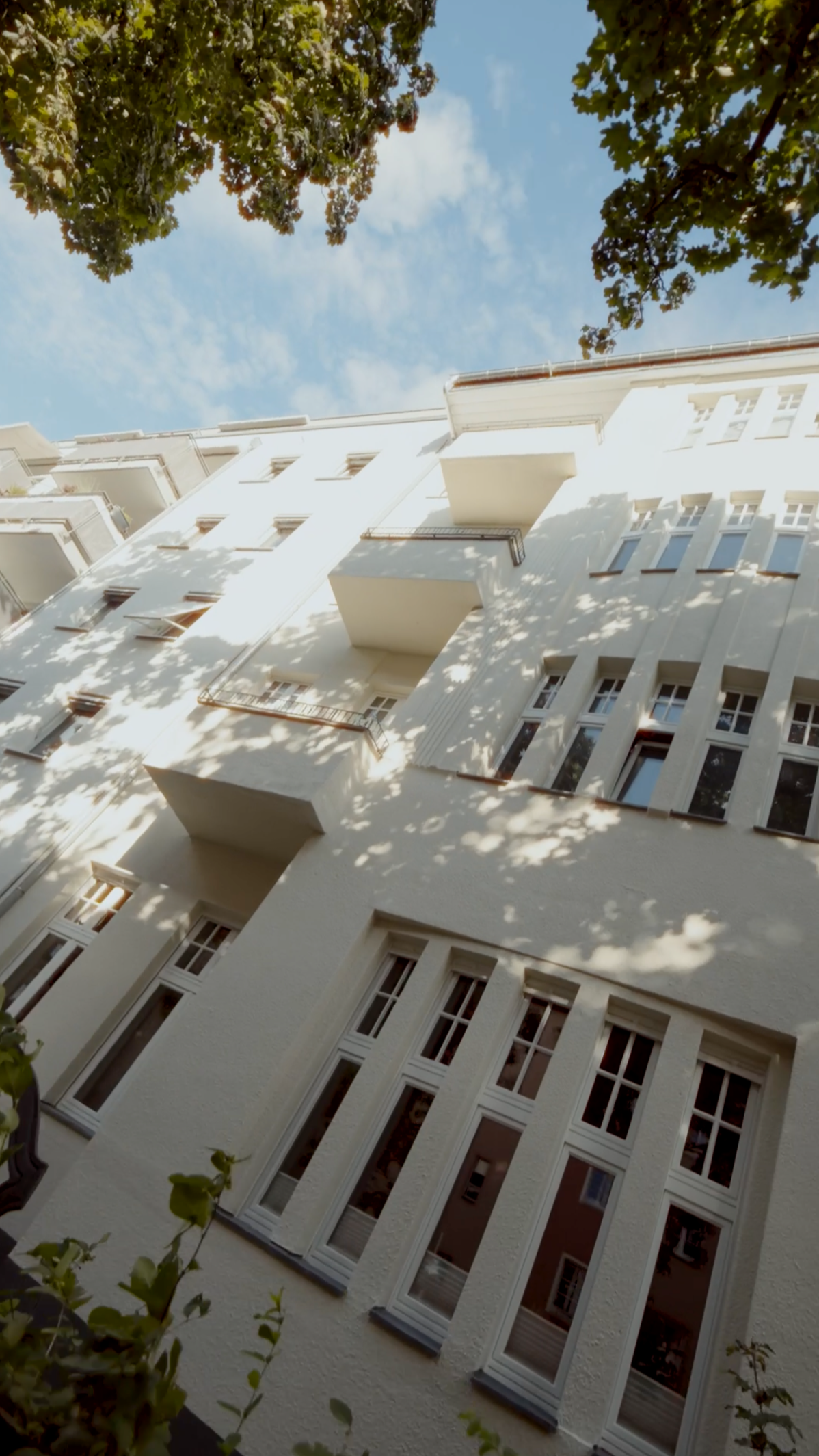Aufnahme eines weißen Wohngebäudes mit mehreren Balkonen, umgeben von Bäumen und einem blauen Himmel mit Wolken