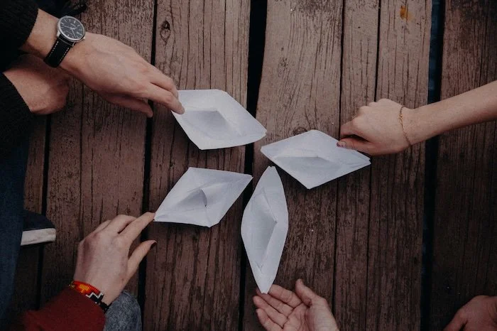 zu sehen sind 4 weiße Papierboote, die in der Mitte zusammenstehen und von 4 verschiedenen Händen in Position gebracht werden. Sie stehen auf einem Holzboden