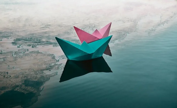 Ein grün-blaues und ein rosa Papierboot auf dem Wasser, sie stehen eng zusammen, das grün-blaue ist vor dem rosafarbenen, sie spiegeln sich im Wasser