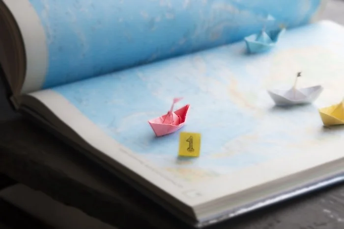 drei Papierboote (pink, blau und weiß) mit Fähnchen auf einem aufgeschlagenen Atlas. Die Papierboote sind an unterschiedlichen Stellen platziert (pink vorne, weiß Mitte, blau hinten). Neben dem pinken Boot ist ein gelbes Schild mit einer 1 drauf