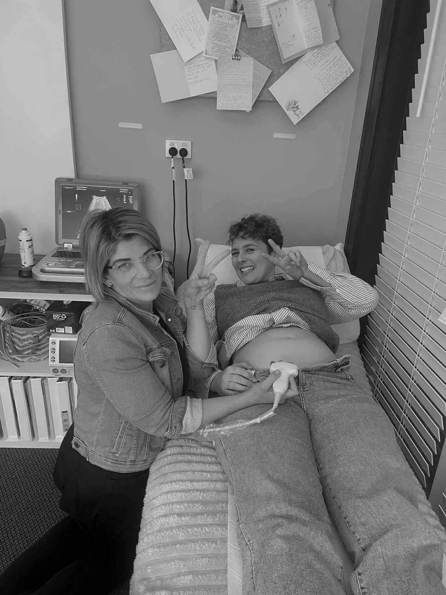 A woman and a pregnant woman in a hospital room, with the pregnant woman lying in bed making peace signs and the woman next to her holding a Doppler ultrasound device, smiling at the camera.