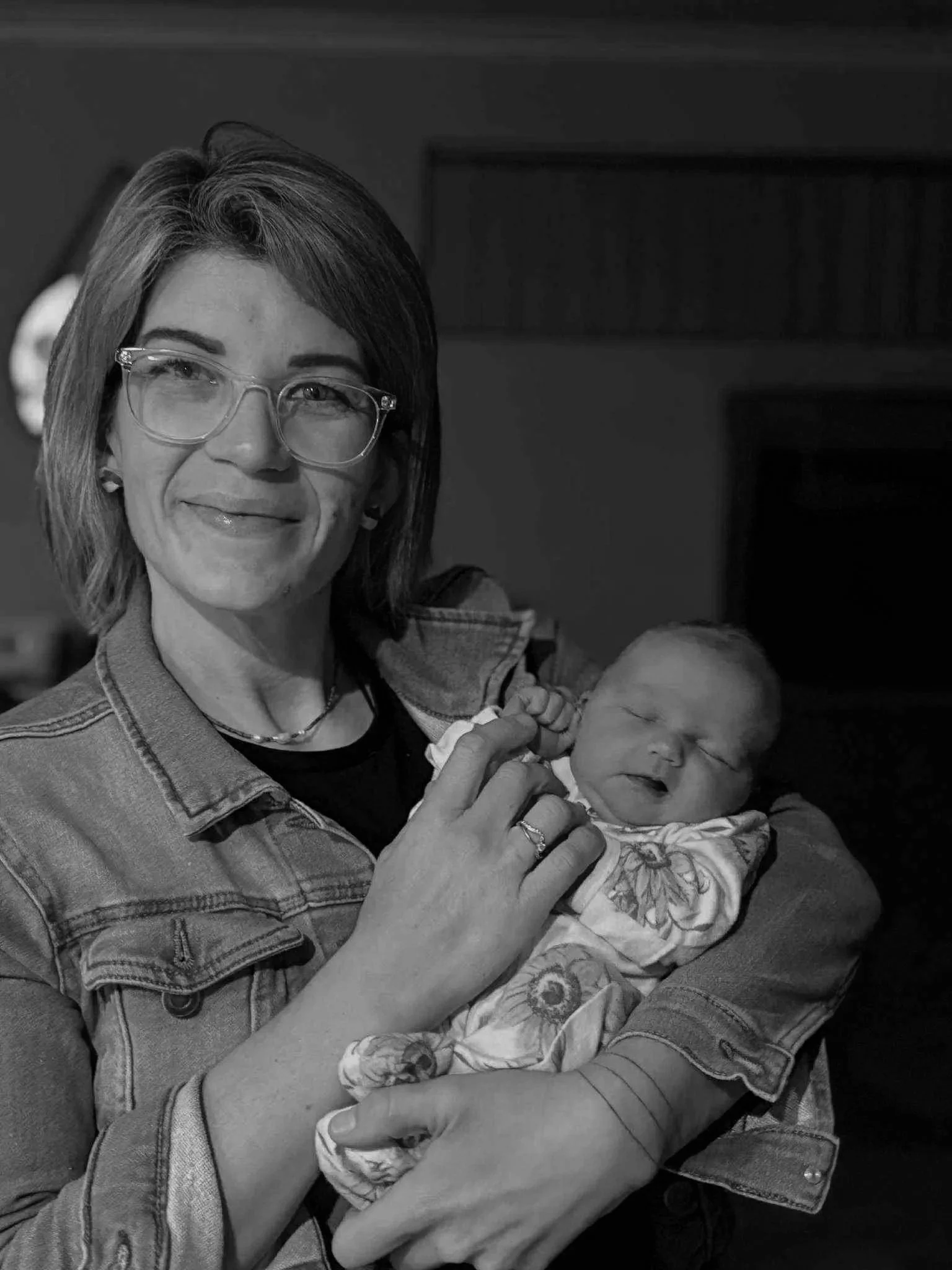 A woman with short hair and glasses holding a smiling sleeping baby in her arms.