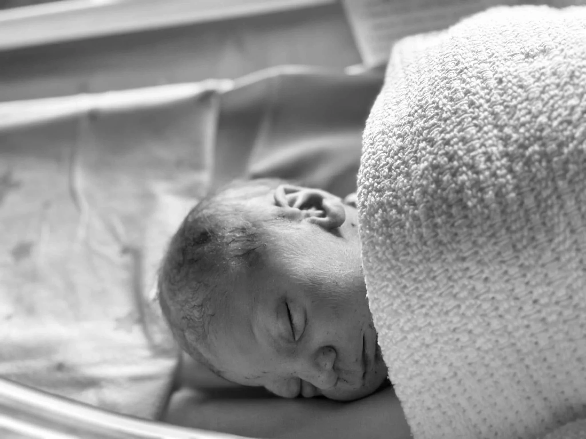 A newborn baby sleeping, wrapped in a towel, lying on a bed or crib with a sheet and a soft textured blanket.