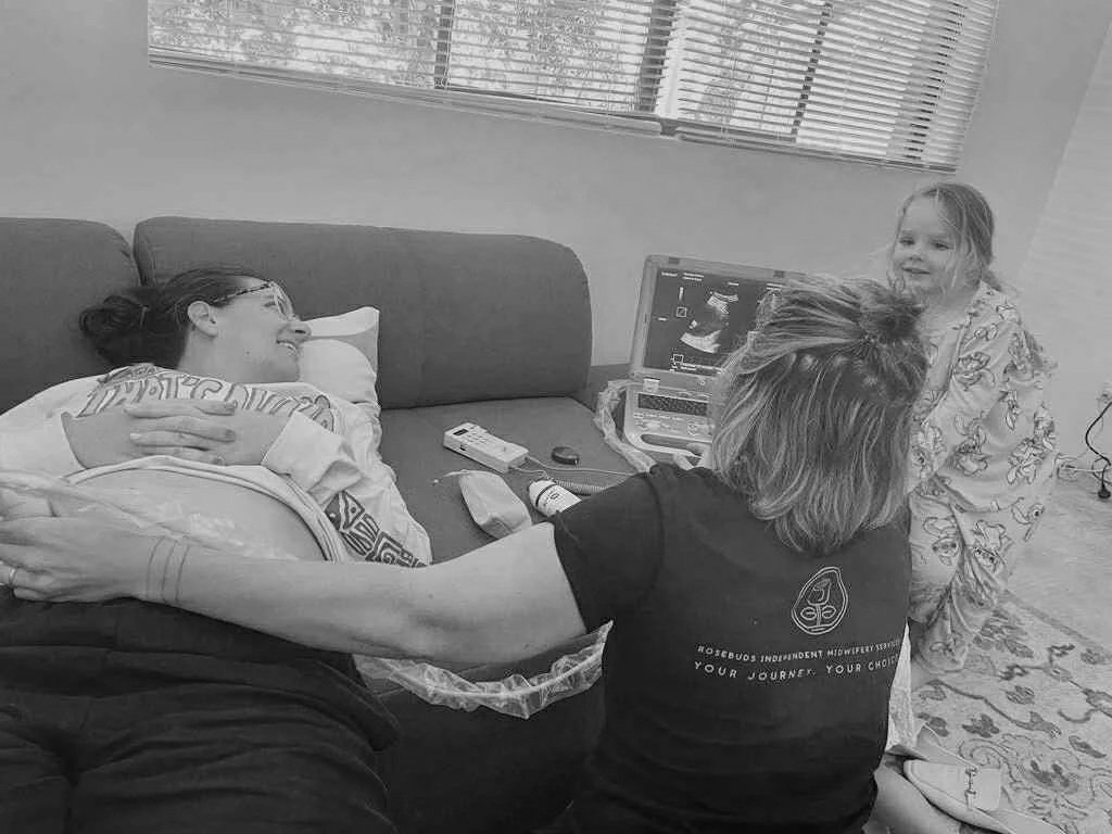 A woman lying on a couch during a hospital ultrasound, with her arm resting on her pregnant belly, while a healthcare professional performs the ultrasound. A young girl in pajamas is standing nearby, smiling at the scene. Medical equipment and an ultrasound monitor are visible.