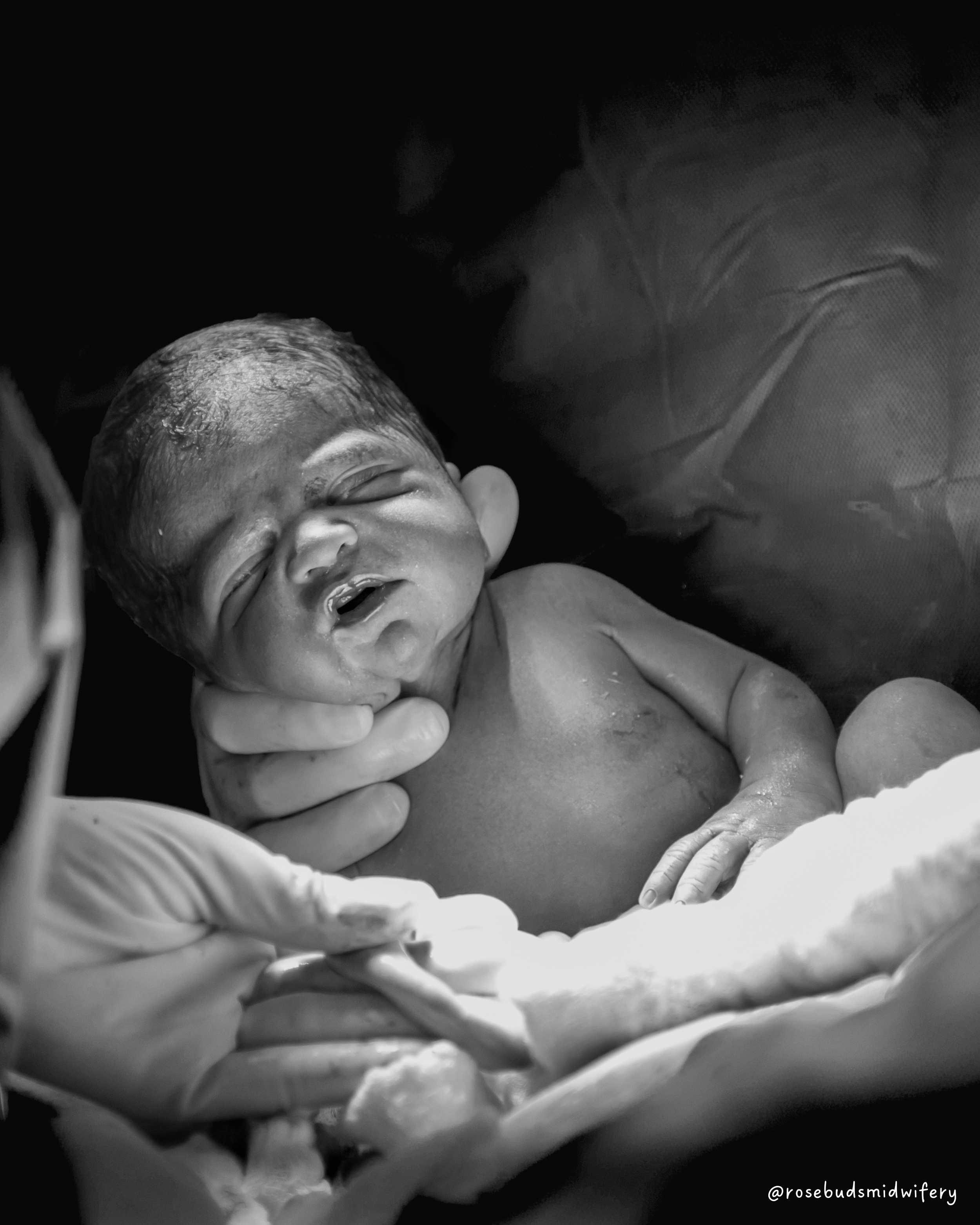 A newborn baby, with eyes closed and mouth slightly open, is being held by a person wearing gloves in a delivery room.