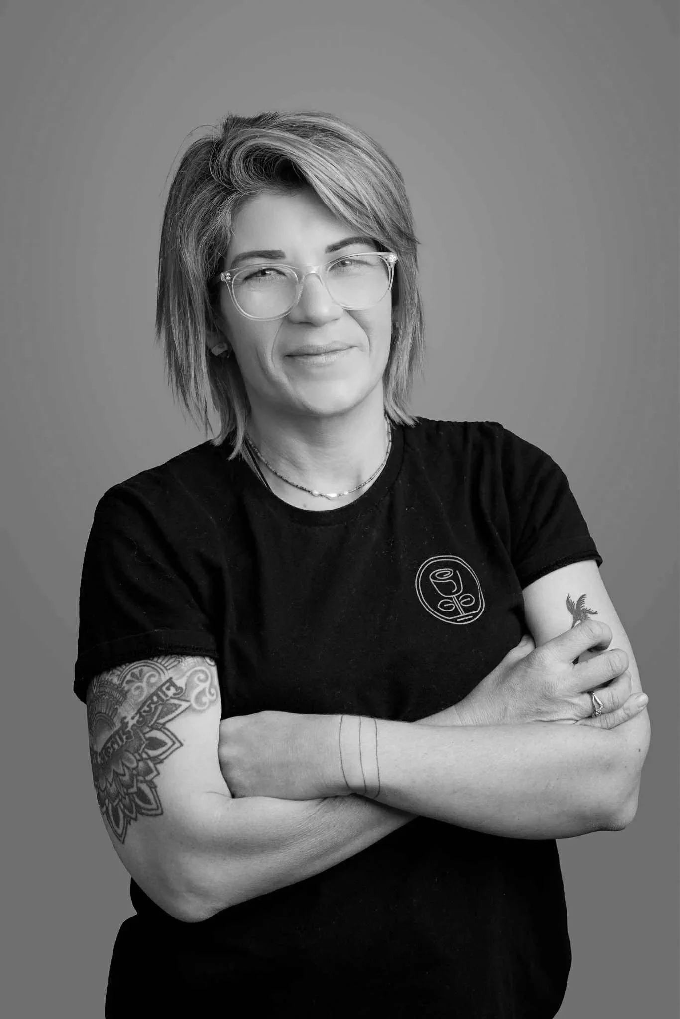 Black and white portrait of a woman with short, layered hair and glasses, crossing her arms, wearing a black t-shirt with a logo, tattoos on her arms, jewelry including necklaces and rings, against a plain background.
