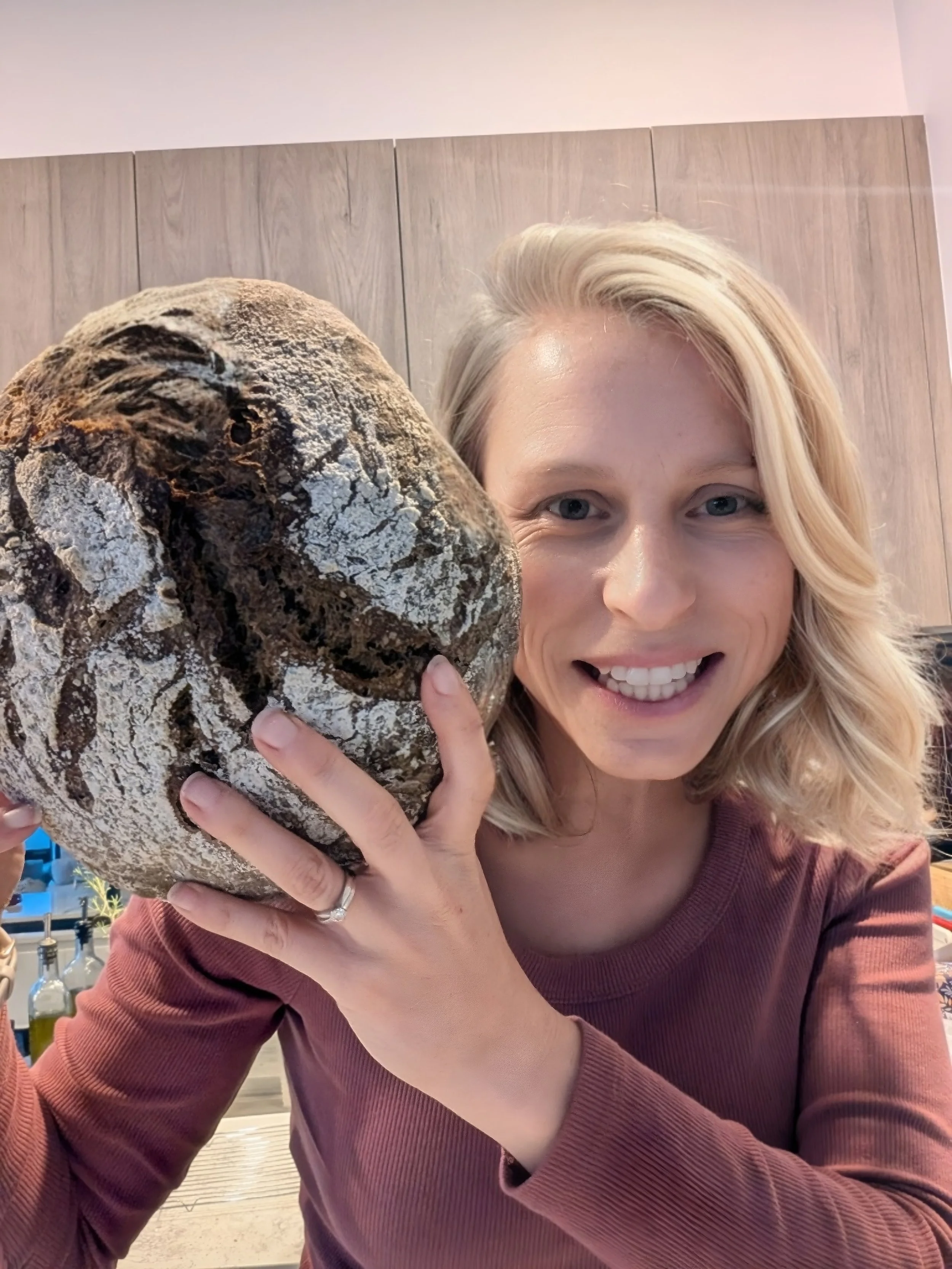 A woman with blonde hair smiling and holding a large, round loaf of bread close to her face in a kitchen.
